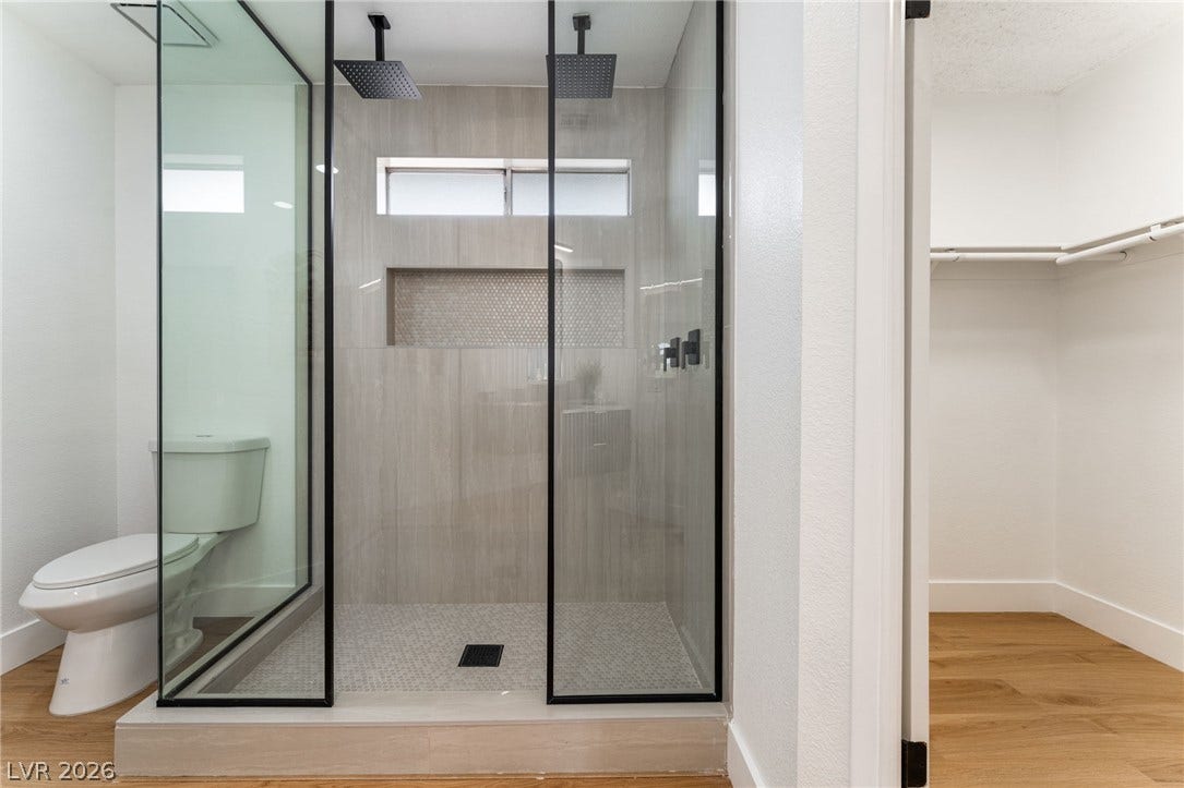 A glass-enclosed walk-in shower features dual showerheads alongside tiled walls and wood flooring.