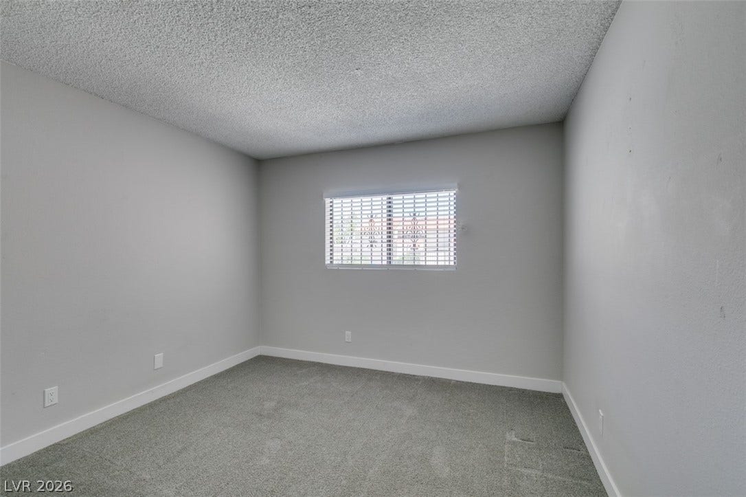 A well-lit bedroom features plush gray carpeting and a wide window with blinds.