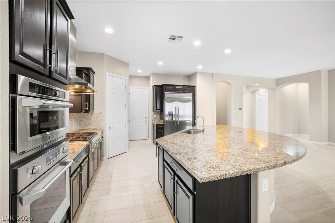 7230 Fresh Harvest Avenue, Las Vegas, NV 89131 A spacious kitchen presents a large island with a sink, elegant dark wood cabinetry, and gleaming Monogram stainless steel appliances.