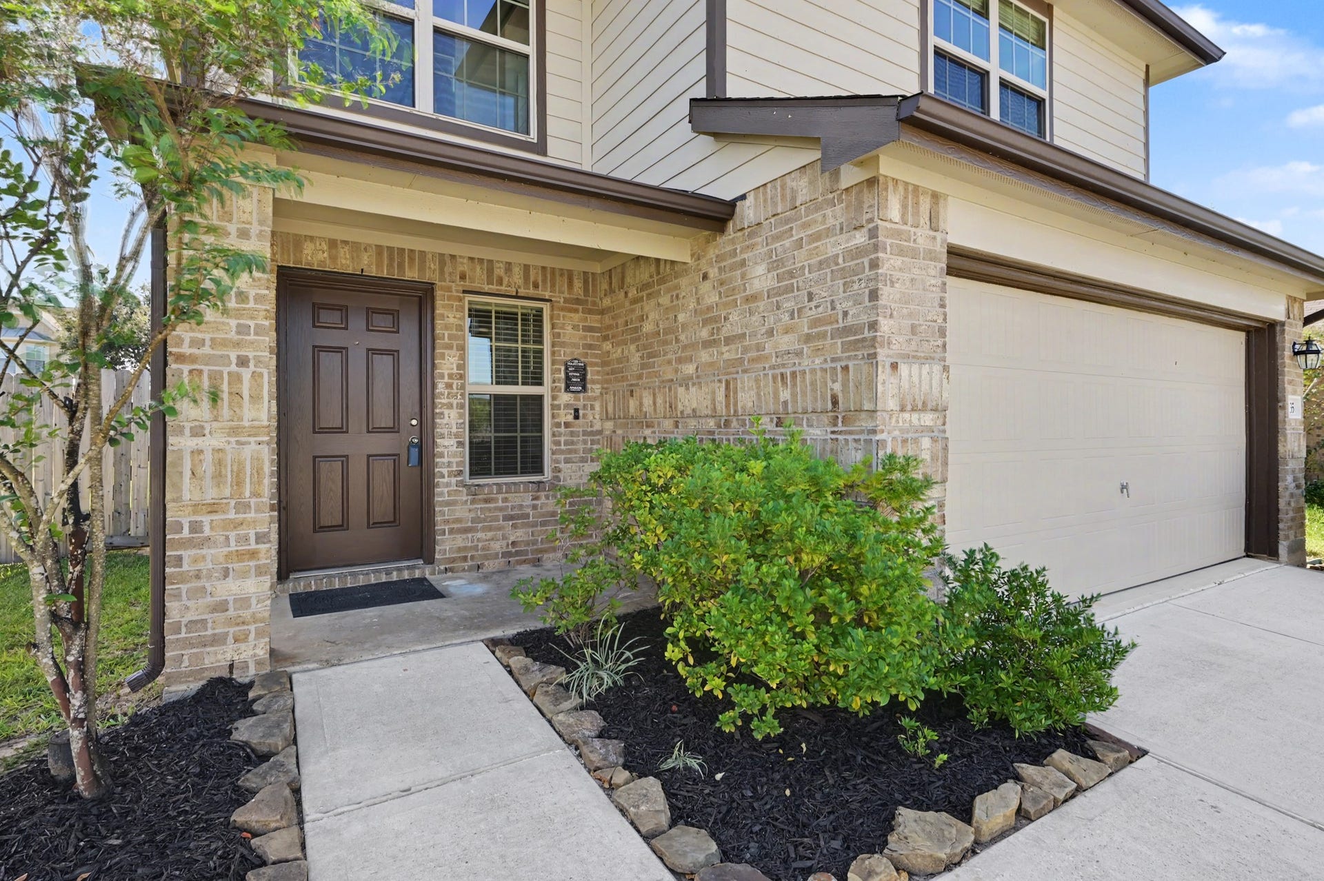 Welcoming covered front entry with attractive brick exterior, beautifully landscaped flower beds, mature greenery, and convenient access to the attached 2-car garage.