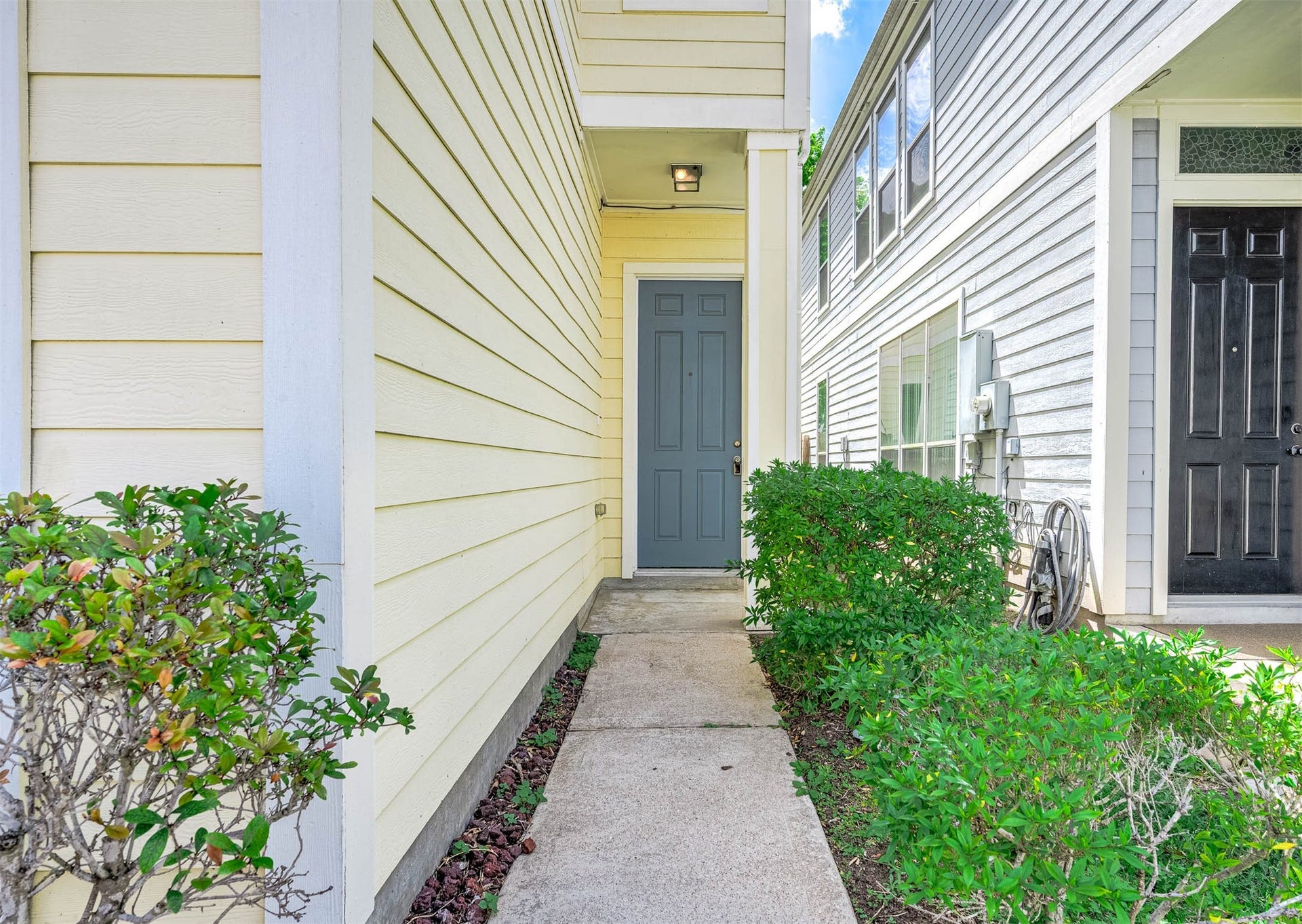 14923 ATMORE Place Dr, Houston, TX 77082 Welcome to your new home, featuring a charming blue door framed by pastel painted siding. Inviting Entrance with Lush green shrubs and grass.