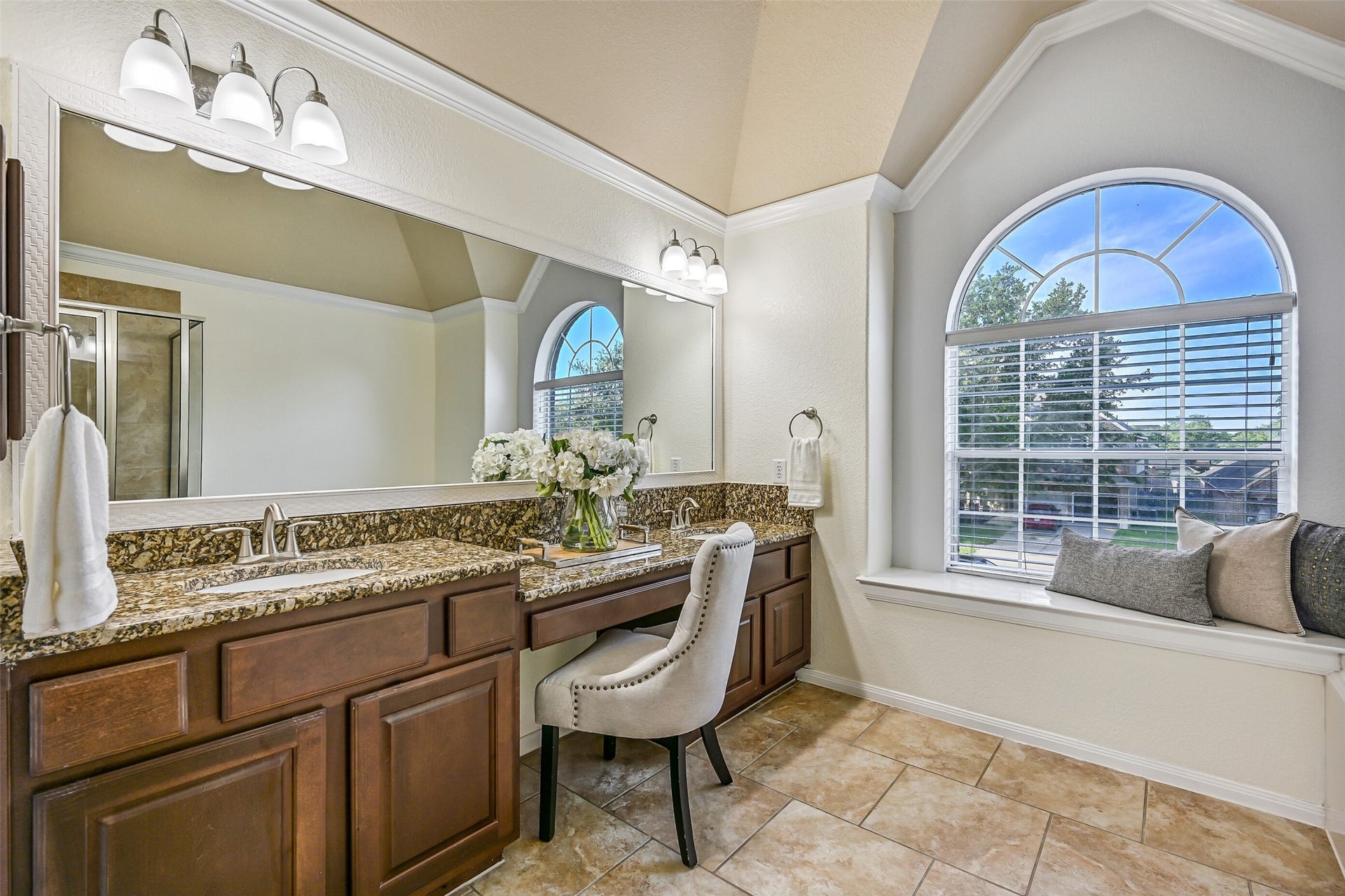 2508 Rockygate Lane, Friendswood, TX 77546 The expansive vanity offers abundant countertop space, handsome raised-panel cabinetry, dual sinks, updated lighting, and a dedicated seated dressing area. The oversized arched window and elevated ceiling details add character and create a bright, airy feel throughout the space.