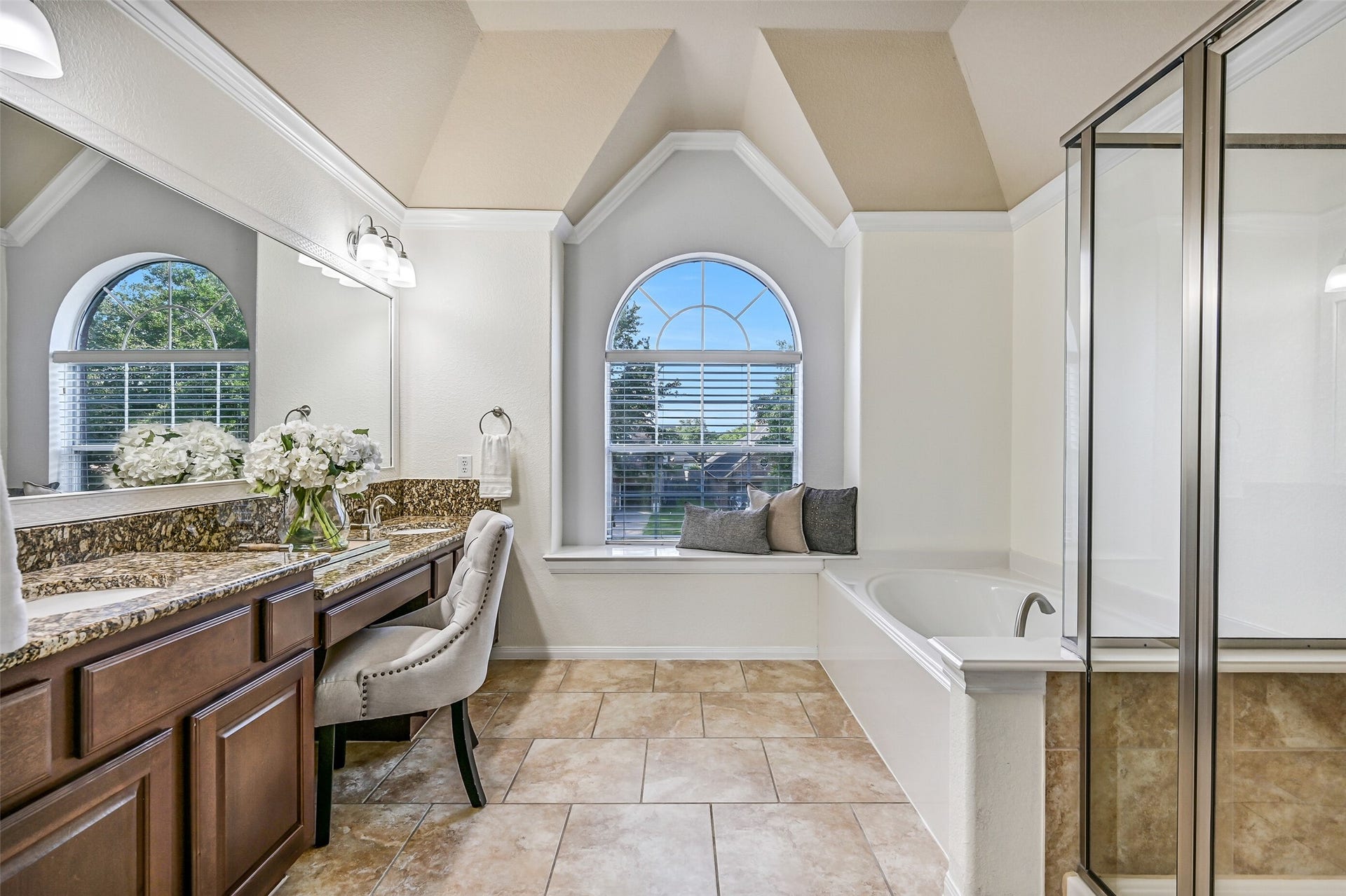 2508 Rockygate Lane, Friendswood, TX 77546 The ensuite bathroom continues the spacious feel of the primary suite with a vaulted ceiling, decorative architectural details, tile flooring, and a granite-topped dual-sink vanity. A built-in seated makeup station sits beneath a large framed mirror, while the arched window fills the room with natural light.