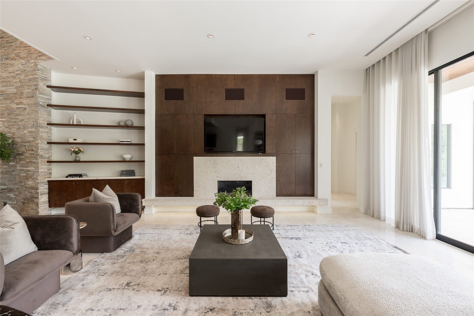 309 Glenwood Drive, Houston, TX 77007 The living room is anchored by a natural travertine fireplace, framed by floating shelves and custom walnut cabinetry that bring warmth and symmetry to the space. Hidden speakers positioned above the cabinetry integrate the surround-sound system discreetly into the architecture, keeping the technology heard but virtually unseen.