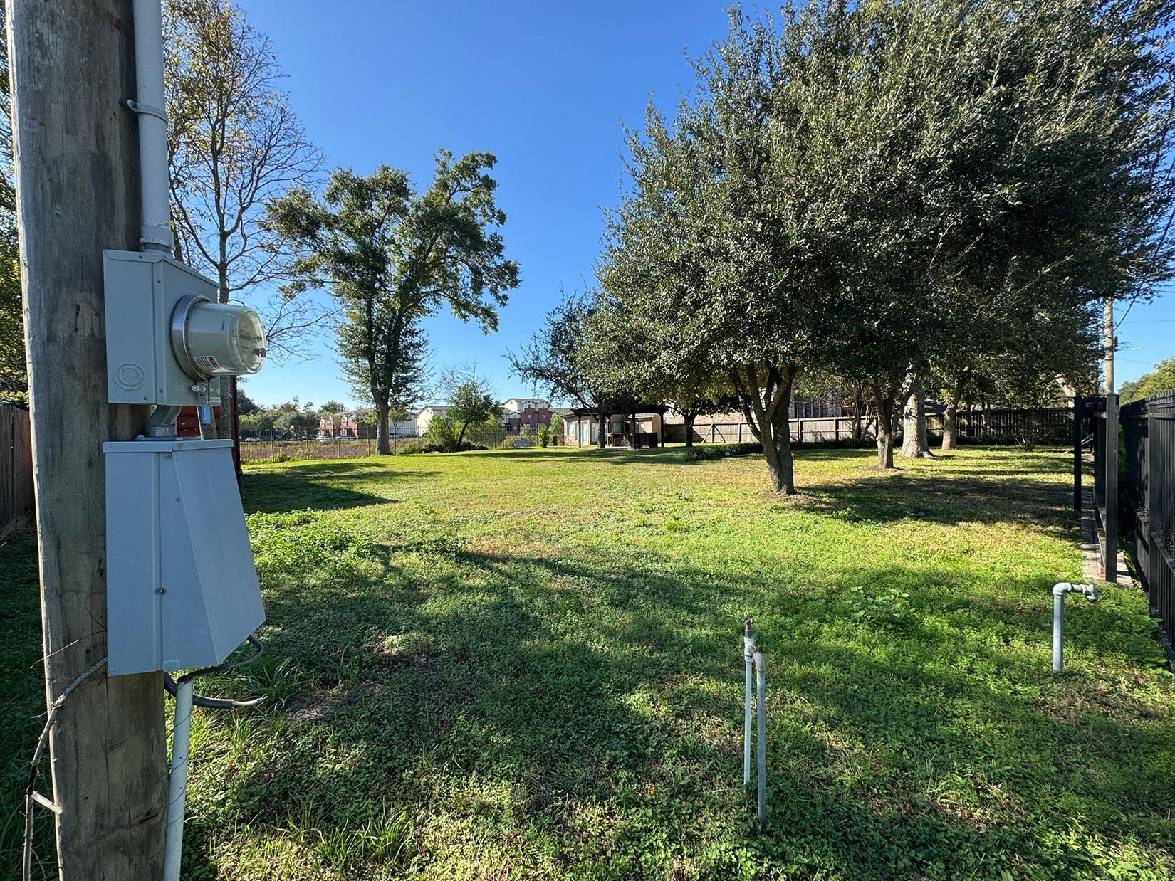 This photo shows a spacious, grassy lot with mature trees, bordered by a fence. Utility connections are present, indicating readiness for development. The area is open and sunny, ideal for building a home.