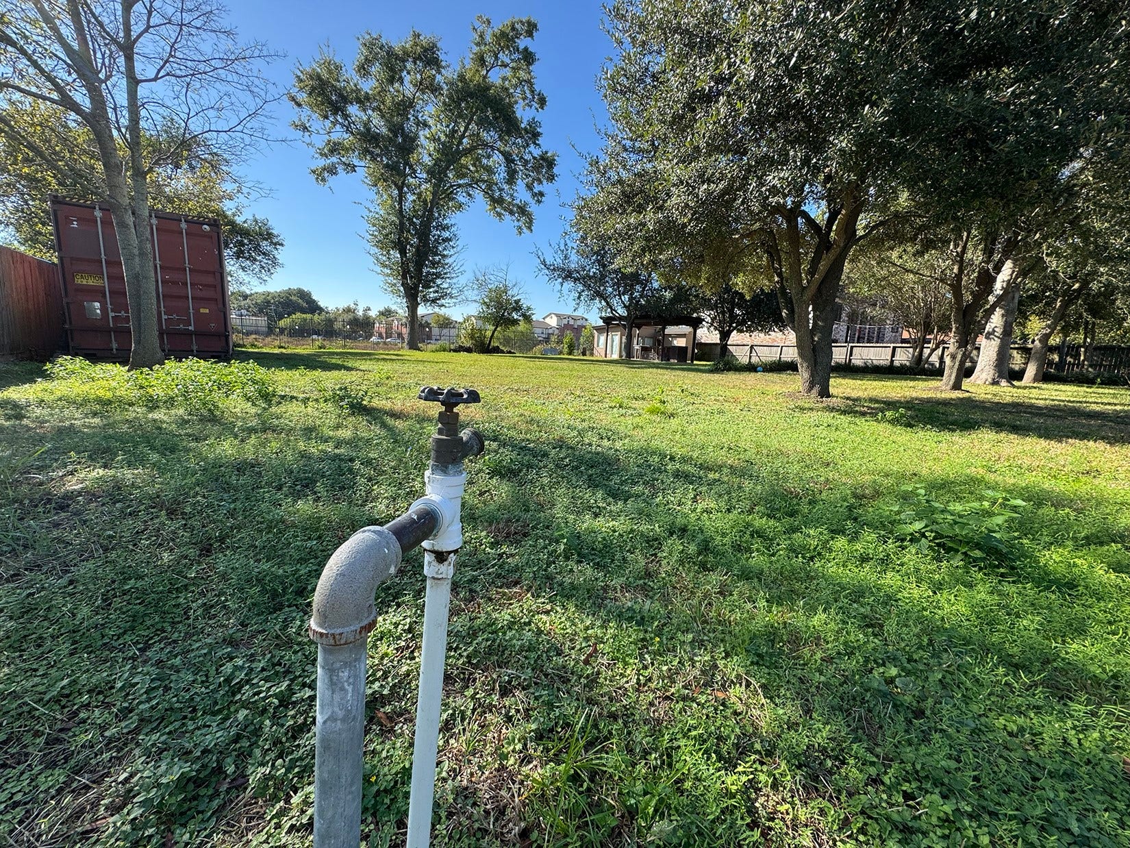Spacious backyard with mature trees, a utility faucet, and a large storage container. The area is sunny and offers potential for landscaping or outdoor activities.
