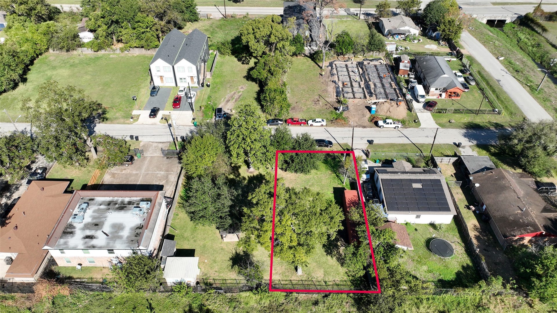 This aerial photo showcases a rectangular lot outlined in red, situated in a residential area. It features ample greenery with trees and open space, surrounded by neighboring homes and buildings. The property is accessible via a paved road, offering potential for development or landscaping.