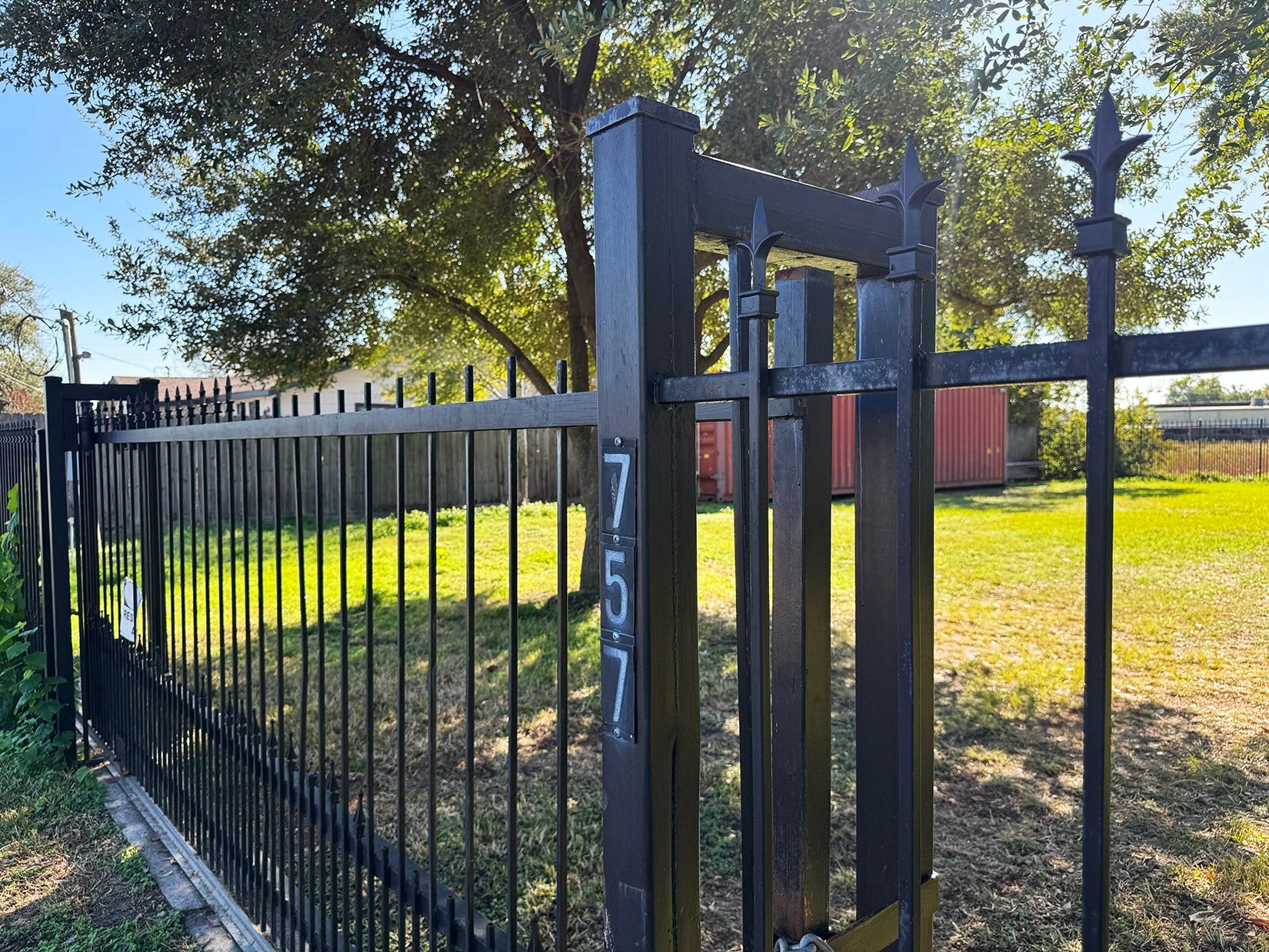 This photo shows a black metal fence with a gate, set in a sunny yard. The area features a well-maintained lawn, mature trees providing shade. Ideal for privacy and outdoor enjoyment.
