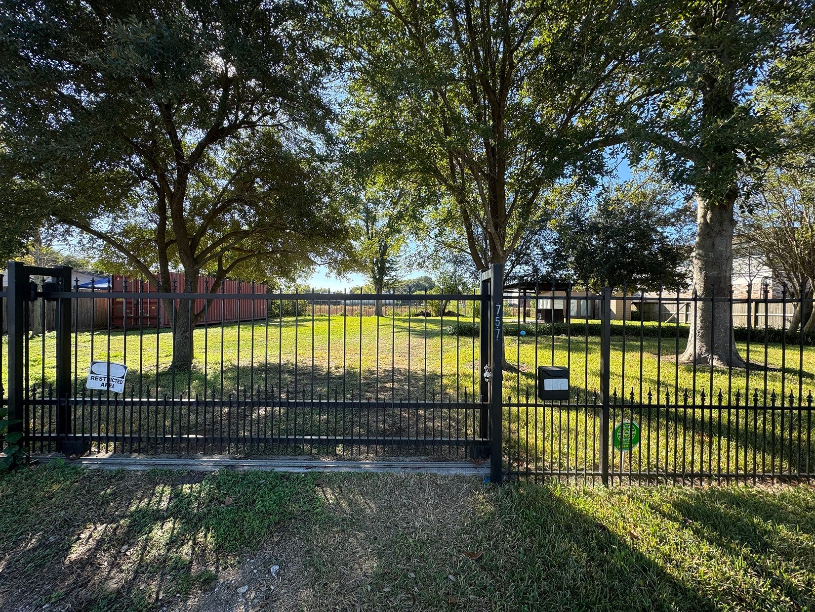 This photo depicts a fenced, grassy lot with mature trees providing shade. The entrance is secured with a black metal gate, offering privacy and a welcoming green space. Ideal for building a home or enjoying outdoor activities.