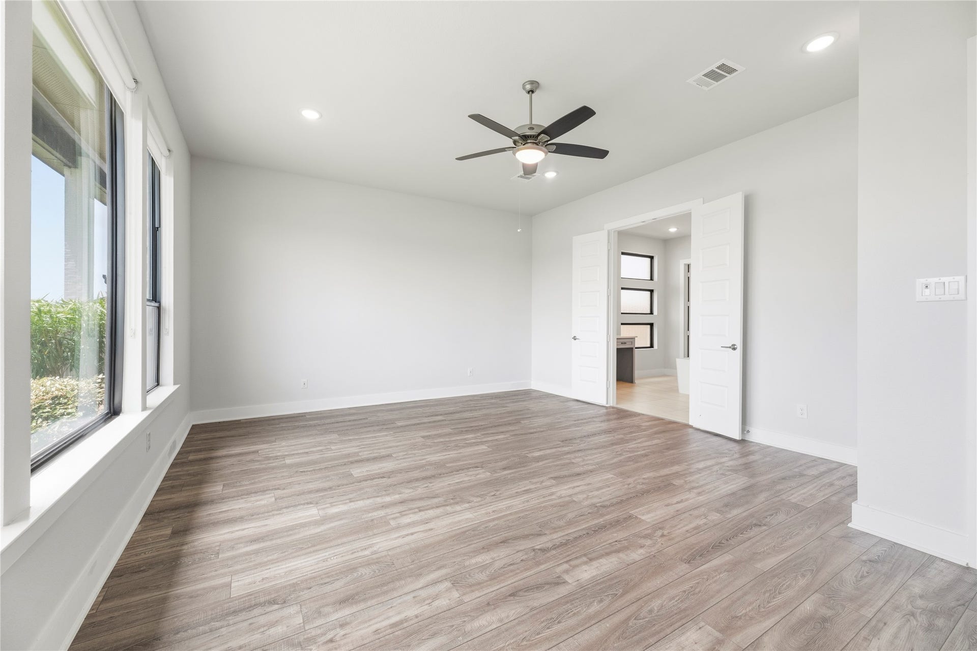 Spacious main-floor primary retreat featuring wall-to-wall wood-look flooring, ceiling fan, large windows, and double doors opening into the primary bathroom.
