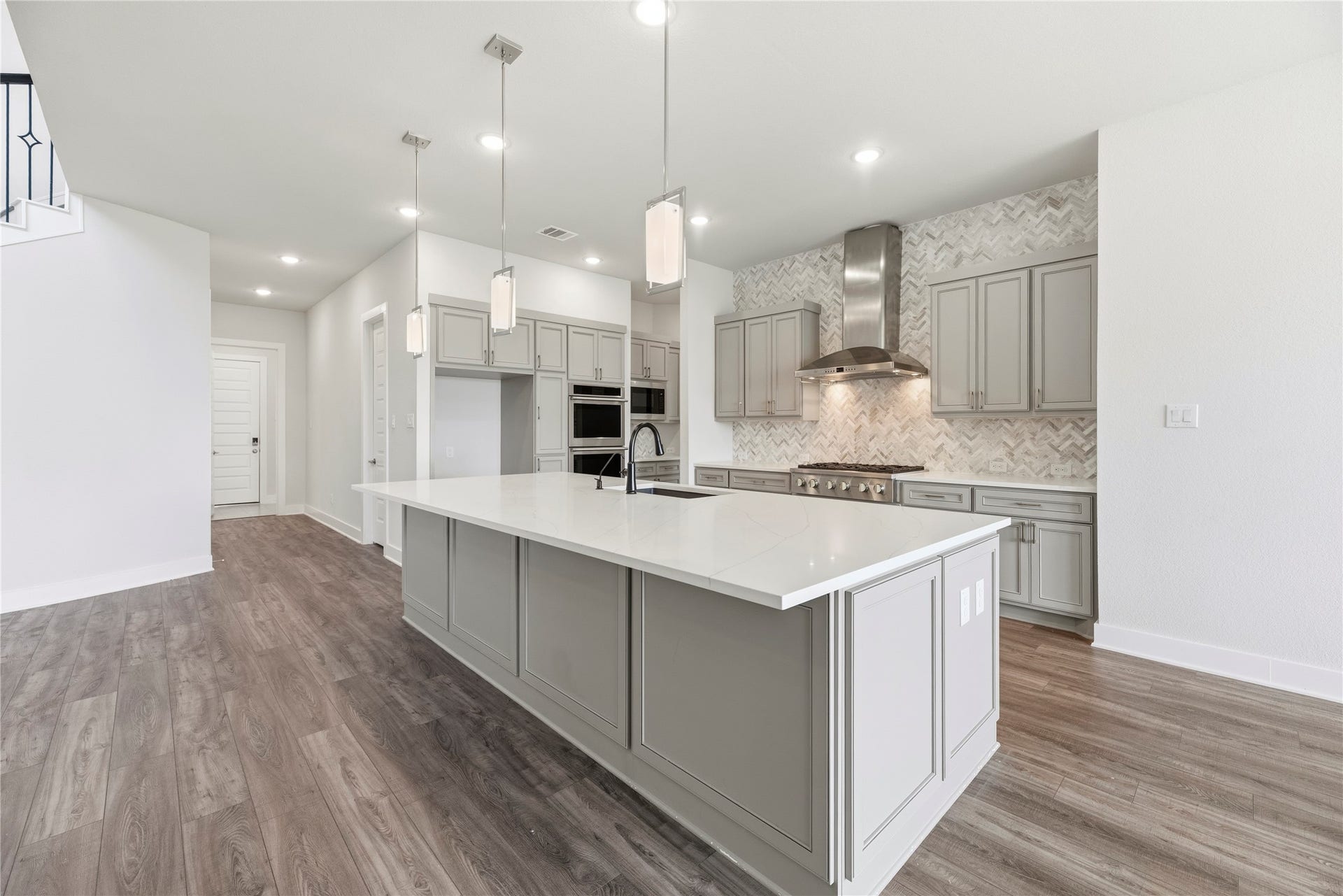 Bright kitchen view highlighting the herringbone tile backsplash, commercial-style gas cooktop, vent hood, double wall ovens, and seamless flow into main living spaces.