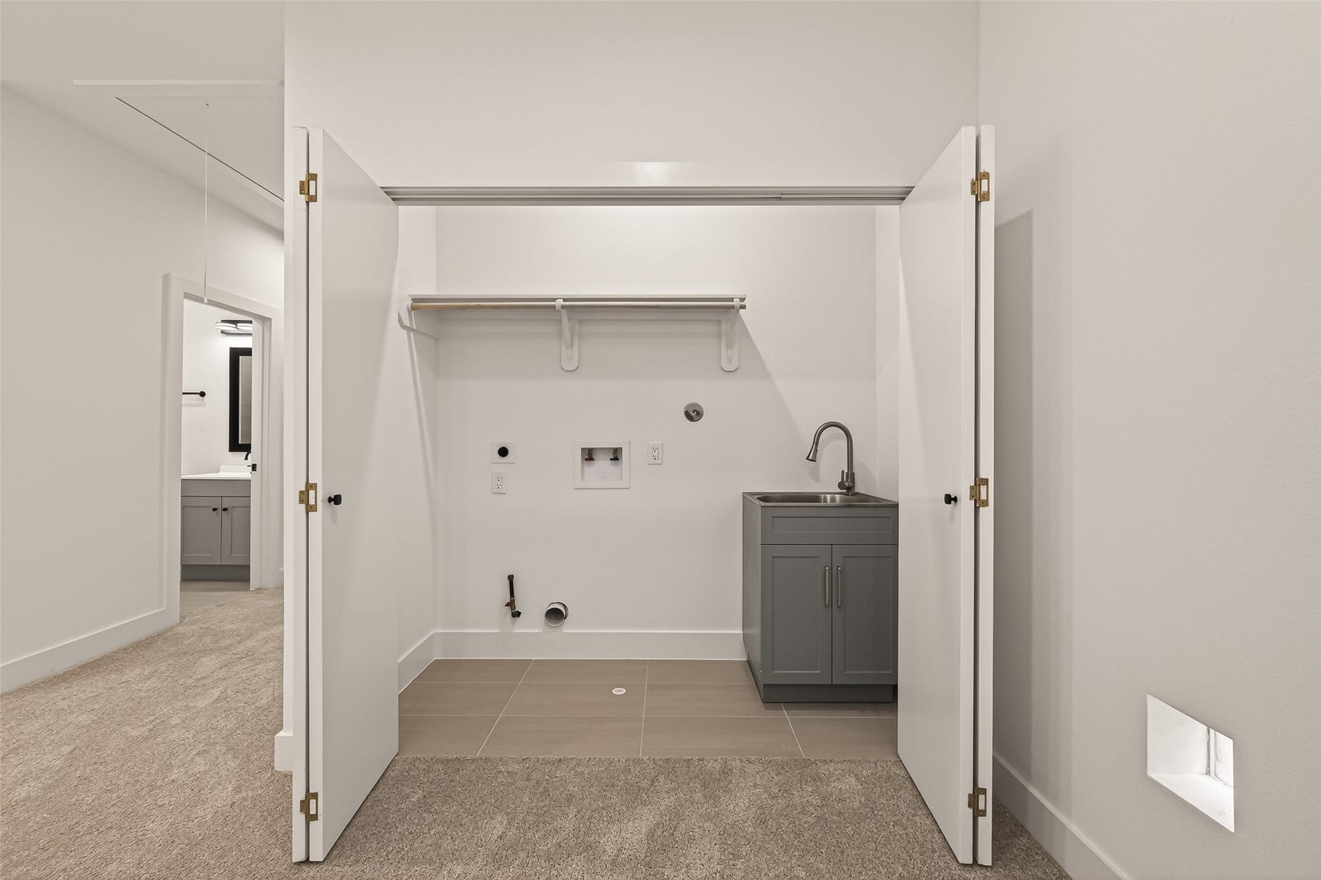 6808 Madrid Street, Houston, TX 77021 This photo shows a laundry area with open double doors revealing hookups for a washer and dryer, a small sink with a cabinet, and a shelf for storage. The room has carpeted flooring leading to a tiled section.