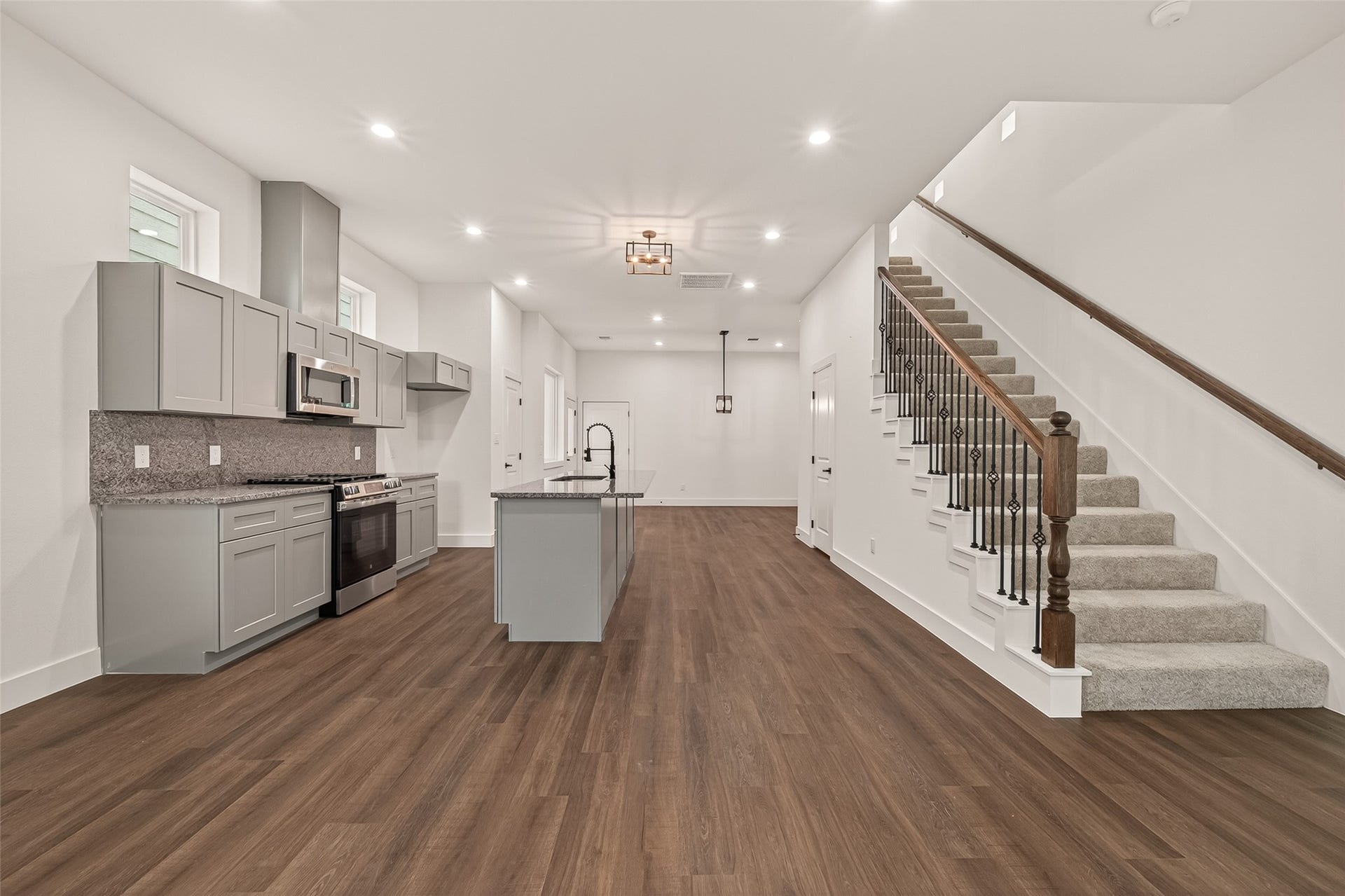 6808 Madrid Street, Houston, TX 77021 This photo showcases a modern, open-concept kitchen and living area with sleek gray cabinetry, stainless steel appliances, and a large island. The space features warm wood flooring and a carpeted staircase with elegant railing, leading to the upper level. Bright and inviting, it offers a spacious setting for family gatherings and entertaining.