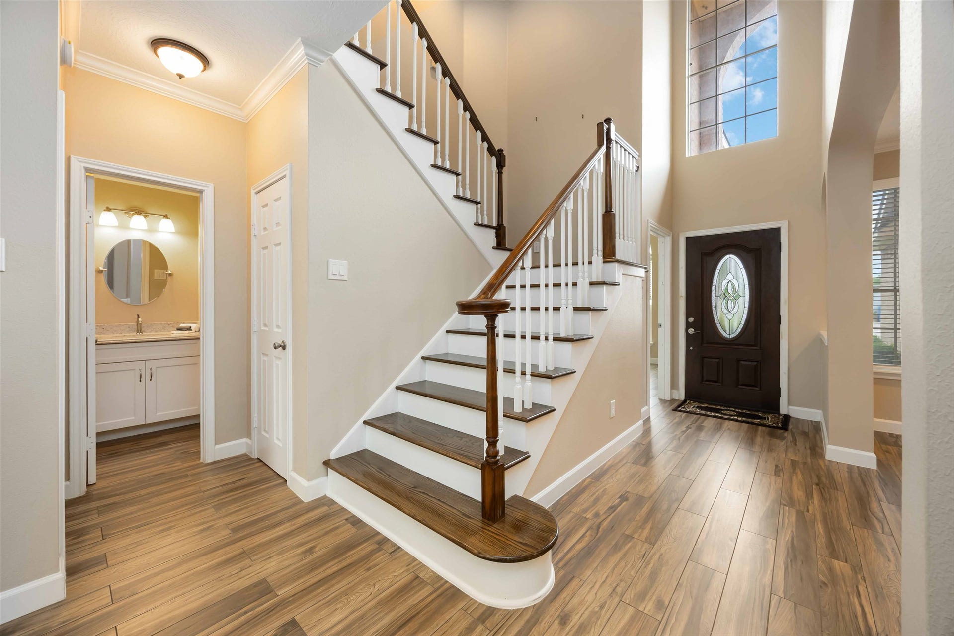 5498 Appleblossom Lane, Friendswood, TX 77546 This photo showcases a bright, welcoming entryway featuring a wooden staircase with white railings, high ceilings, and a large window. The space includes wood-look flooring and leads to a bathroom with a modern vanity.