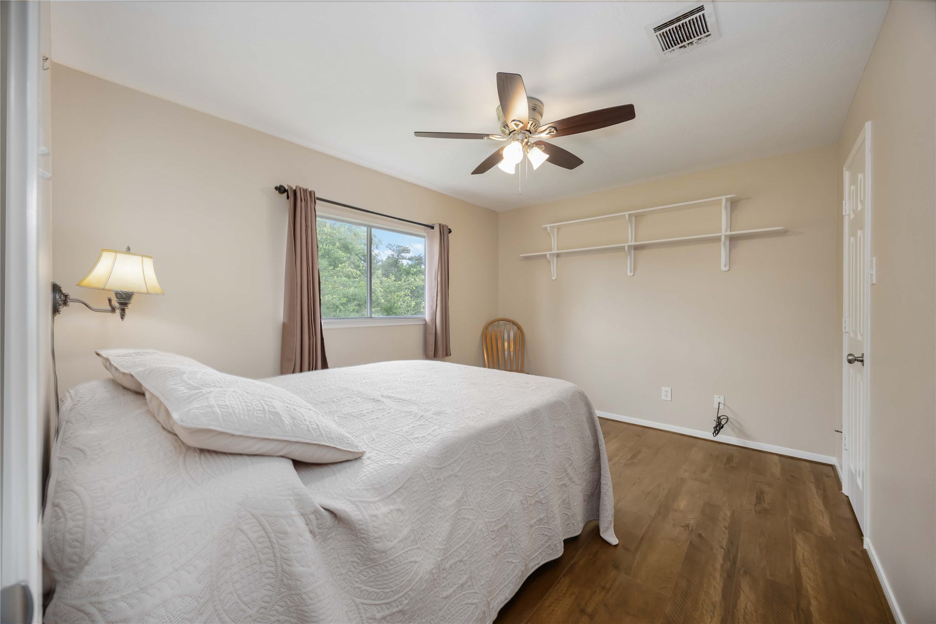 5498 Appleblossom Lane, Friendswood, TX 77546 This bedroom features a cozy, neutral-toned space with a ceiling fan, wood flooring, and a large window with a view. It includes a wall-mounted lamp, shelf for storage, and ample natural light.