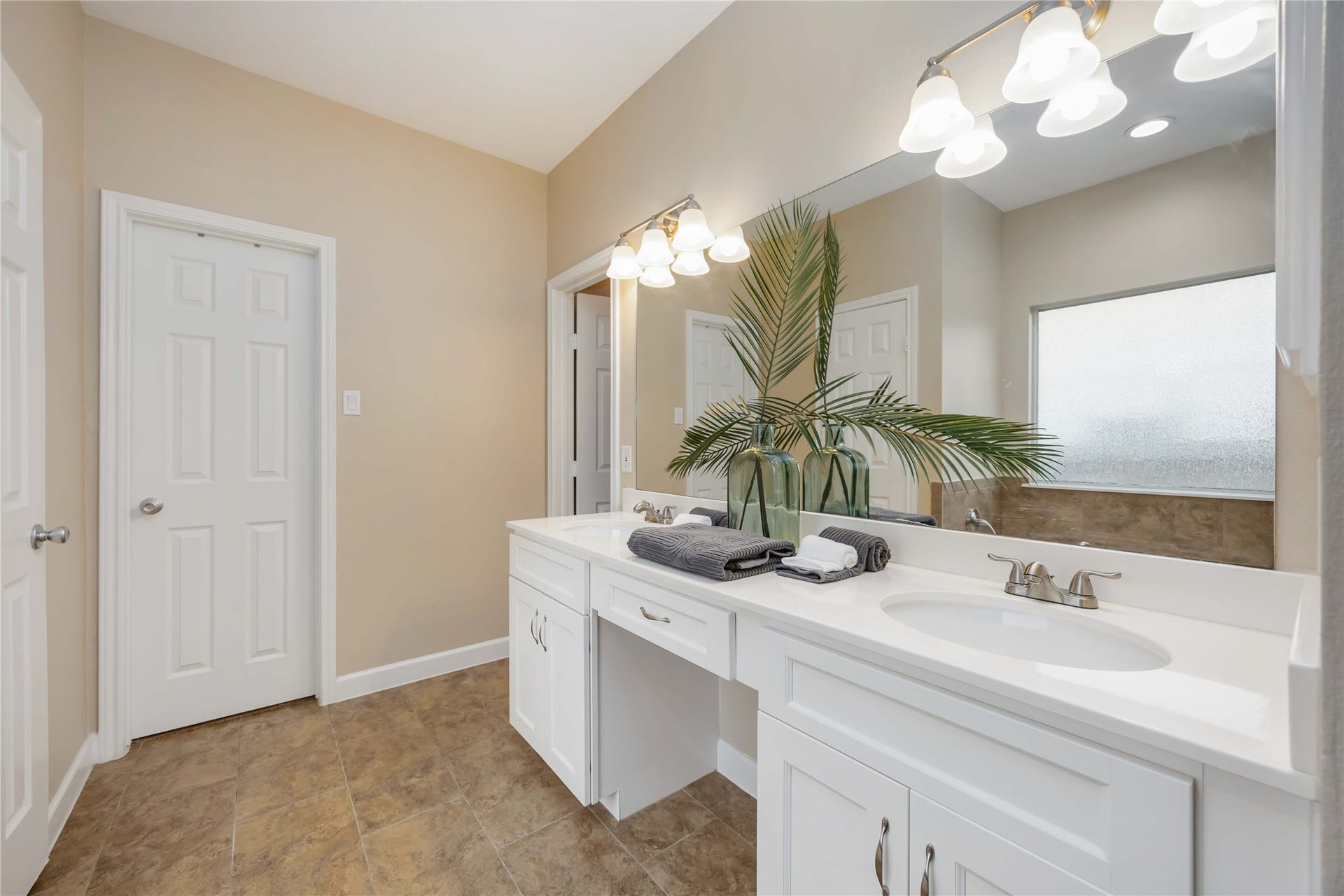 5498 Appleblossom Lane, Friendswood, TX 77546 Bright and spacious bathroom with a large mirror, dual sinks, and modern lighting. It features elegant cabinetry, a frosted window for privacy, and a decorative plant for a fresh touch.
