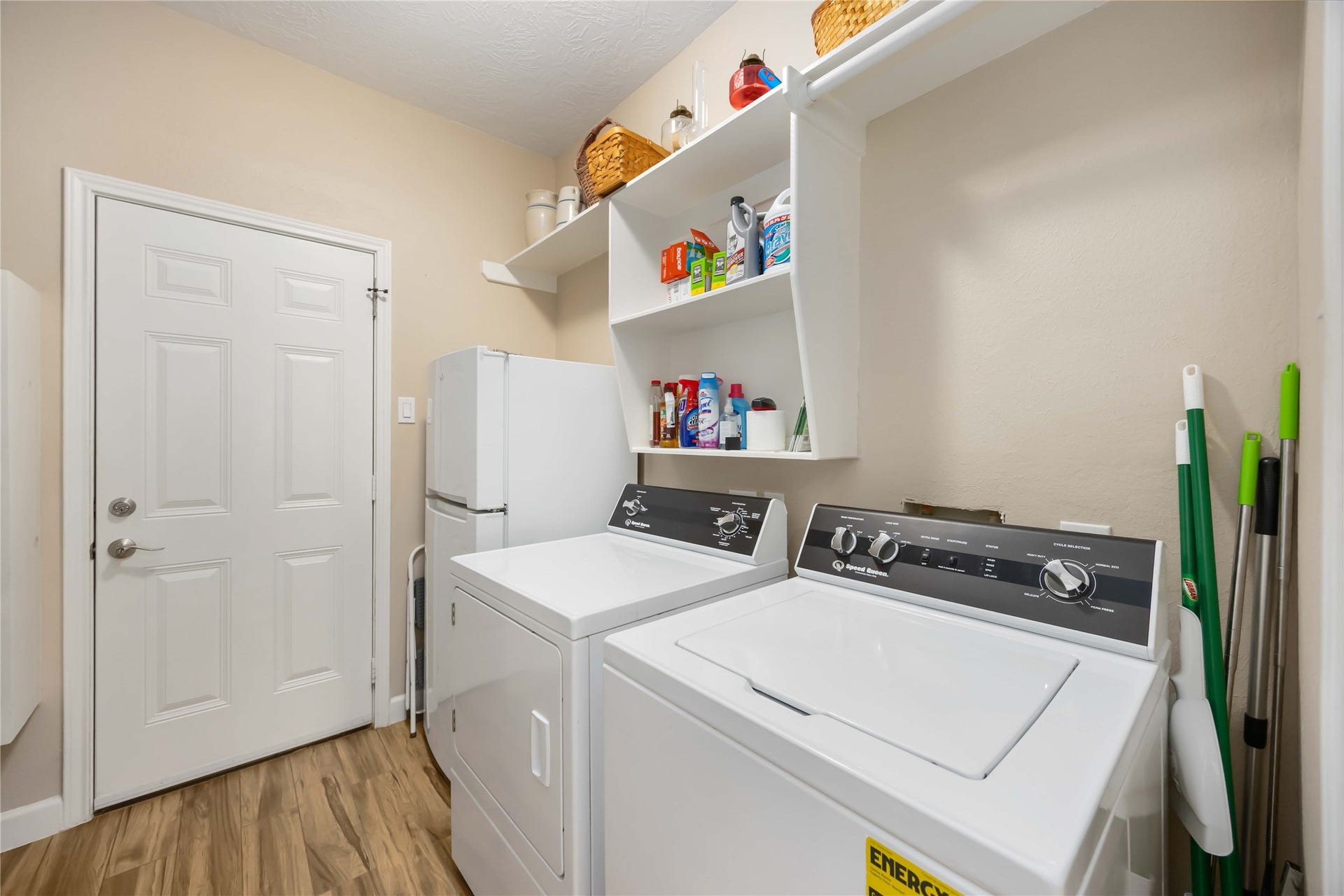 5498 Appleblossom Lane, Friendswood, TX 77546 This laundry room with a washer and dryer, a white storage shelf above for cleaning supplies, and a utility closet. The room has a clean, modern look with wood-style flooring and neutral walls.
