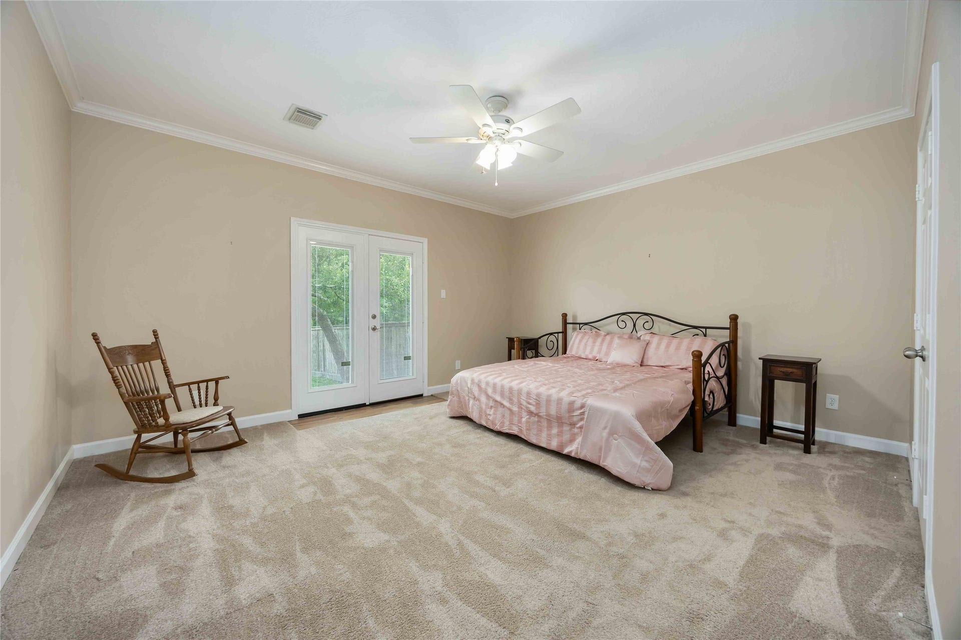 5498 Appleblossom Lane, Friendswood, TX 77546 Spacious primary bedroom with neutral walls and carpet, featuring a ceiling fan for comfort. It includes a decorative bed and side tables, a rocking chair, and French doors leading to an outdoor space.