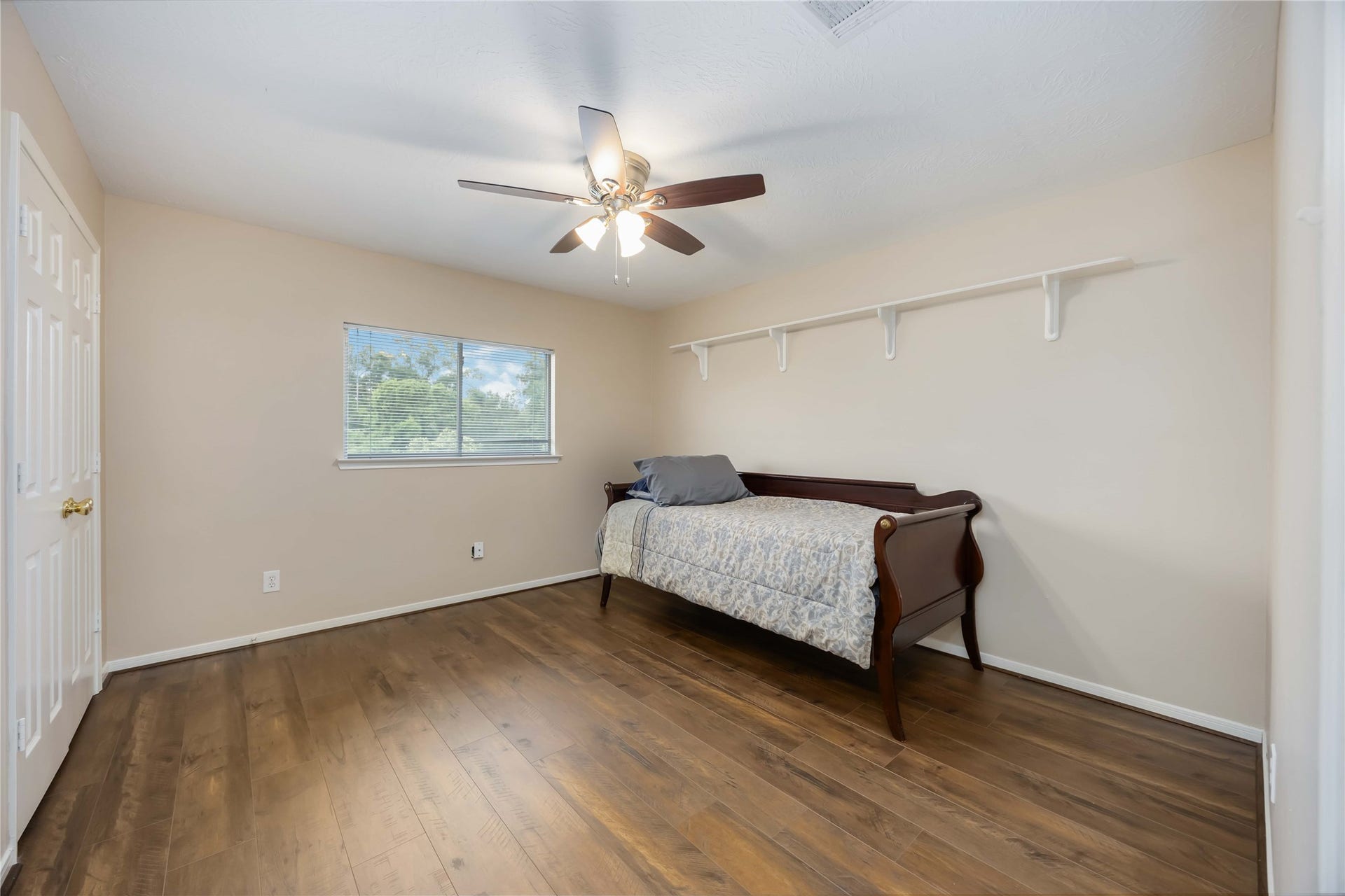 5498 Appleblossom Lane, Friendswood, TX 77546 This room features a warm wood floor, neutral walls, and a ceiling fan. A window provides natural light, and a shelf offers extra storage. It's currently furnished with a daybed, making it versatile for a bedroom or office.