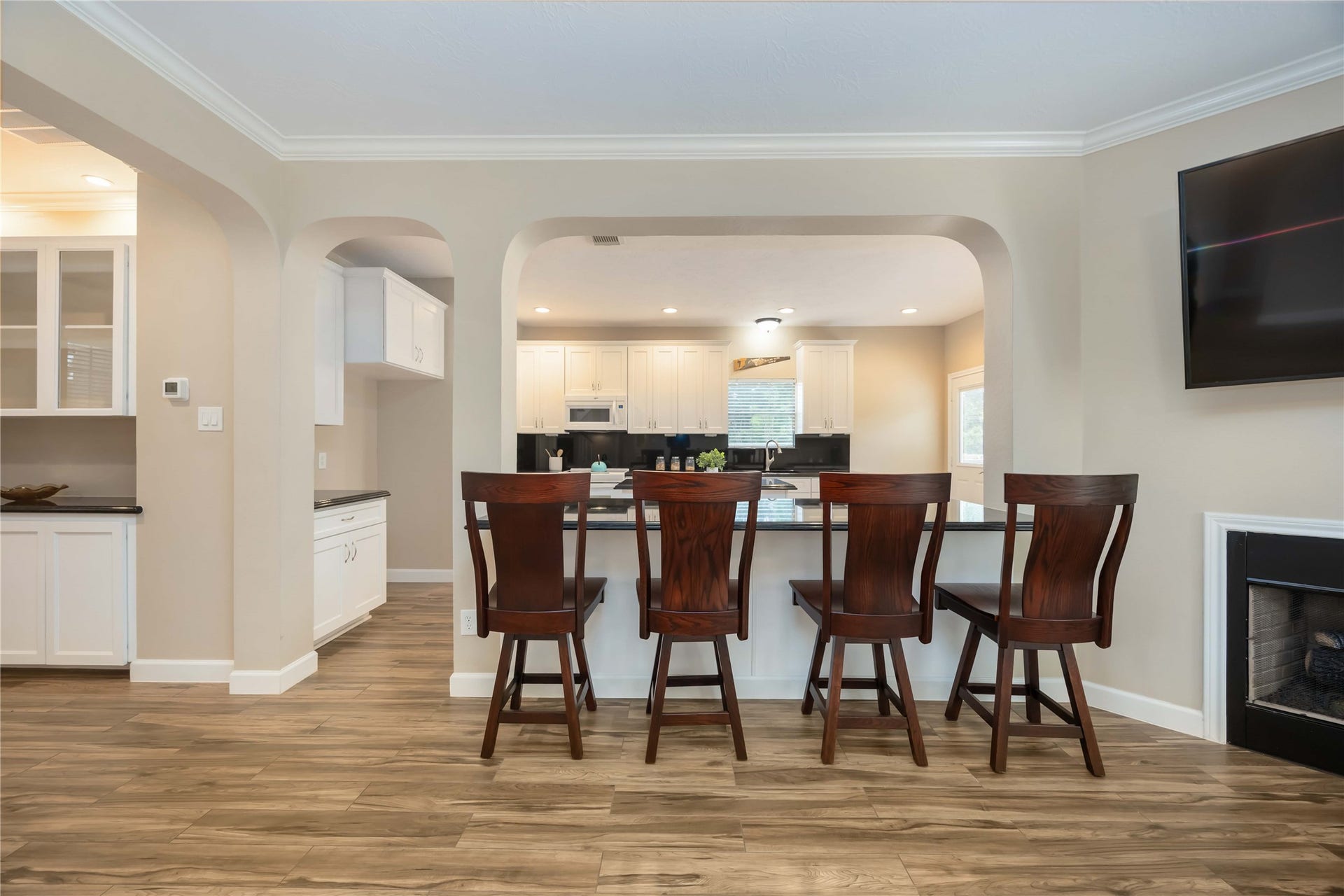 5498 Appleblossom Lane, Friendswood, TX 77546 An open-concept kitchen with a breakfast bar featuring four wooden stools. The space is bright with modern white cabinetry, a sleek countertop, and a visible living area with a fireplace. The flooring is a warm wood finish, enhancing the cozy, inviting atmosphere.