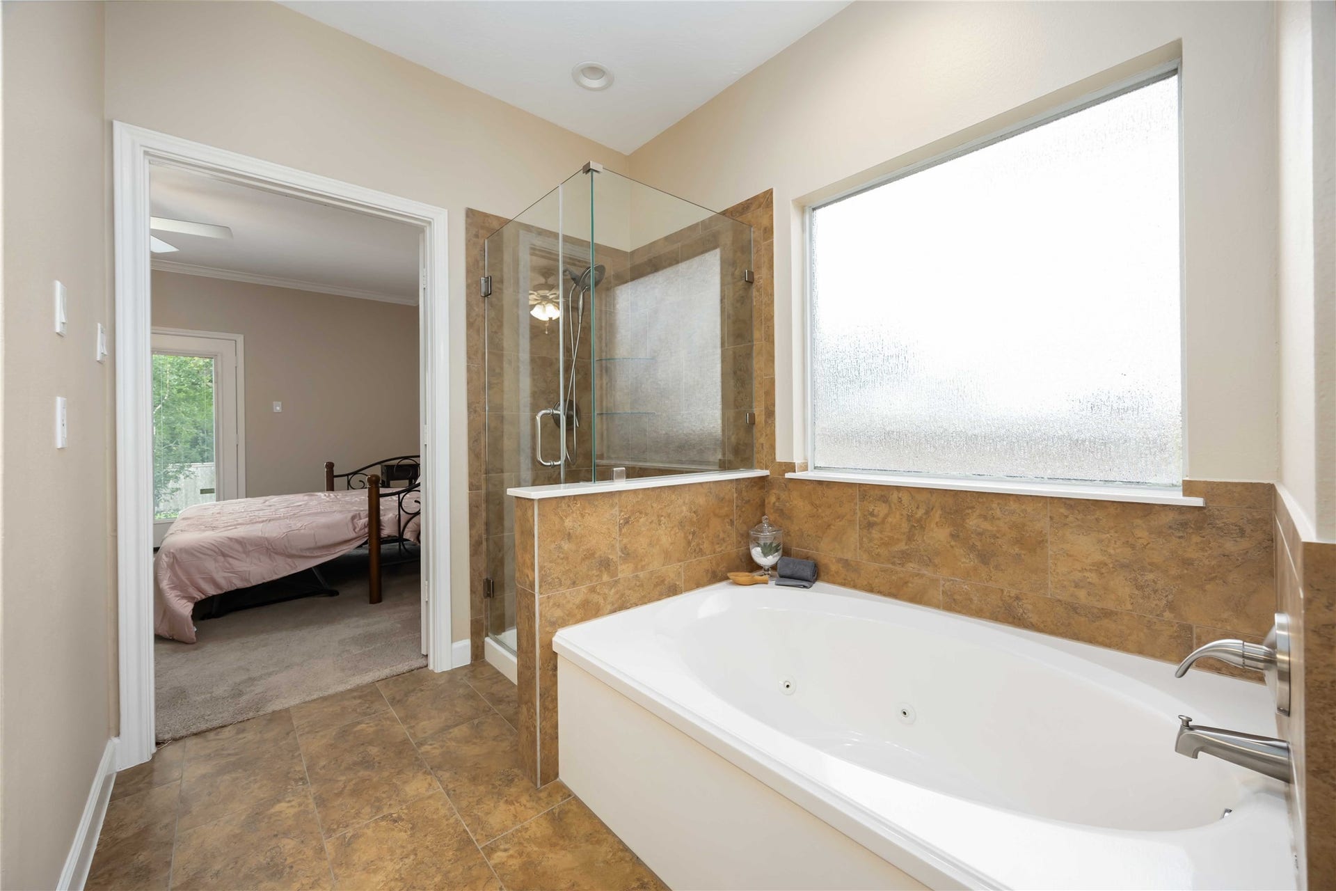 5498 Appleblossom Lane, Friendswood, TX 77546 Bright, modern bathroom with a spacious bathtub, glass-enclosed shower, and a large frosted window for natural light. It opens into a cozy bedroom with a view of greenery outside.