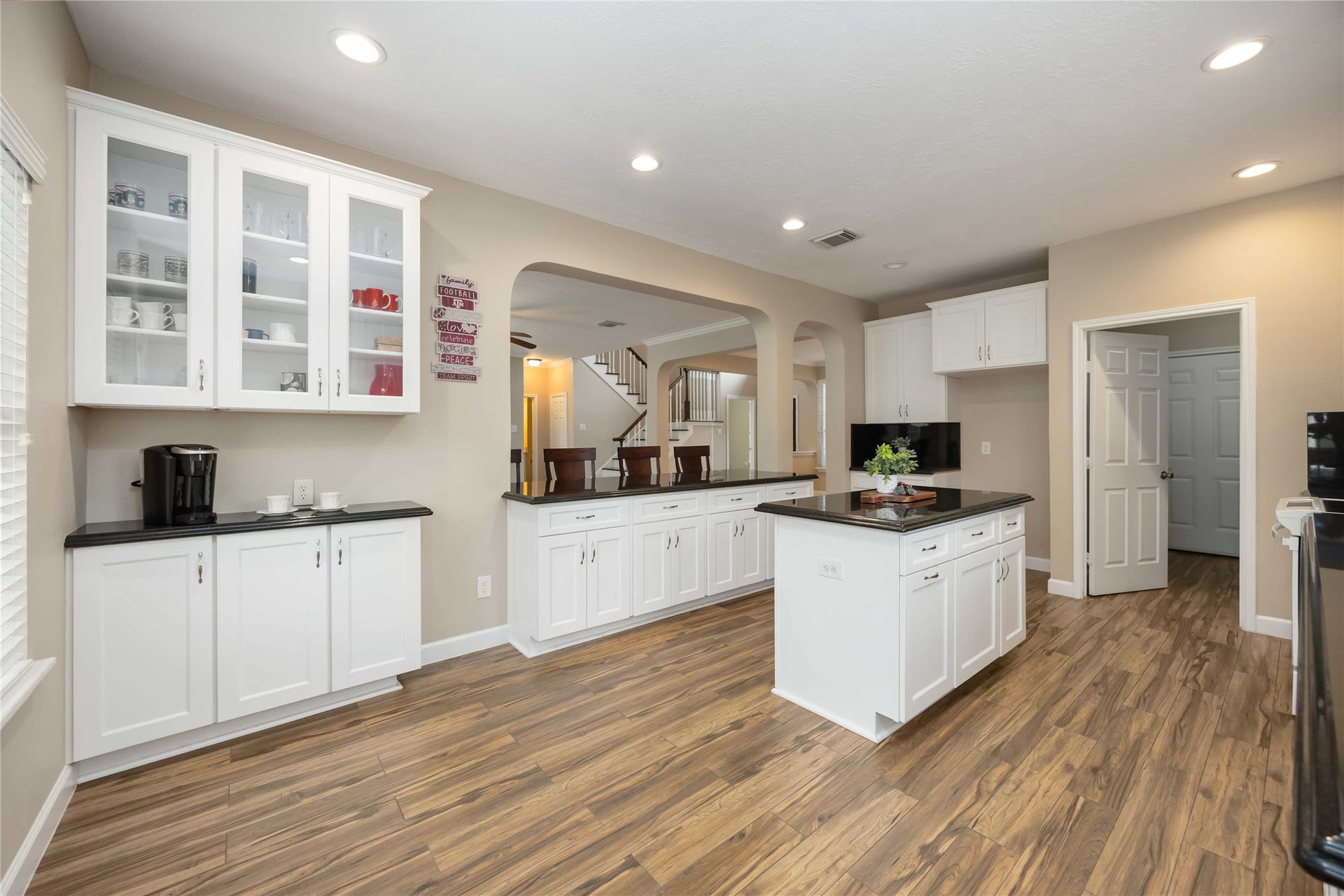 5498 Appleblossom Lane, Friendswood, TX 77546 This kitchen features a modern design with hardwood floors, white cabinetry, and dark countertops. It includes a central island, recessed lighting, and open shelving, creating a bright and welcoming space. The layout offers easy access to adjacent rooms through open archways.