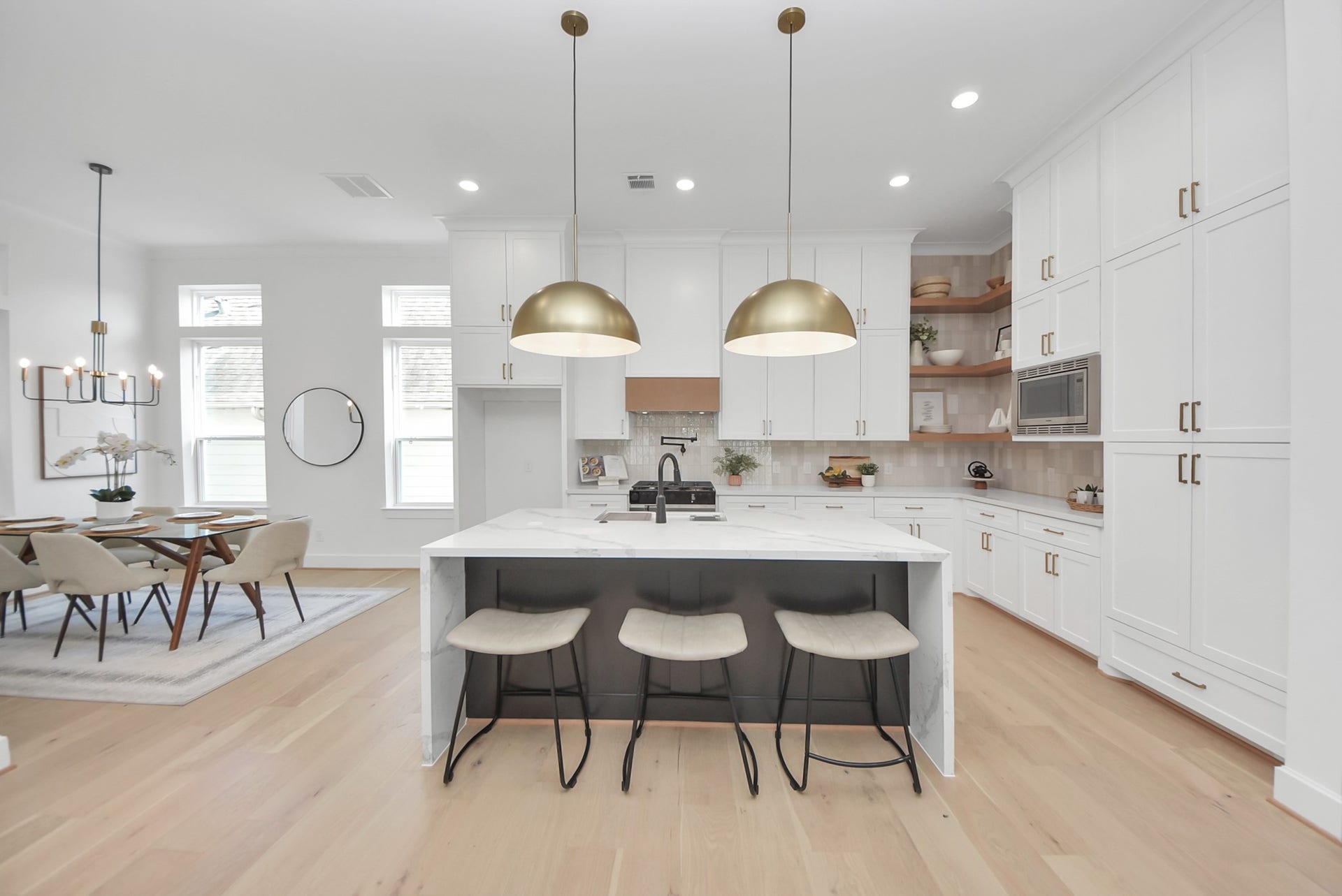 923 Fisher Street, #E, Houston, TX 77018 This modern kitchen features elegant white cabinetry, a large marble island with seating, and stylish pendant lighting. The open layout flows into a dining area with ample natural light, creating a bright and inviting space.