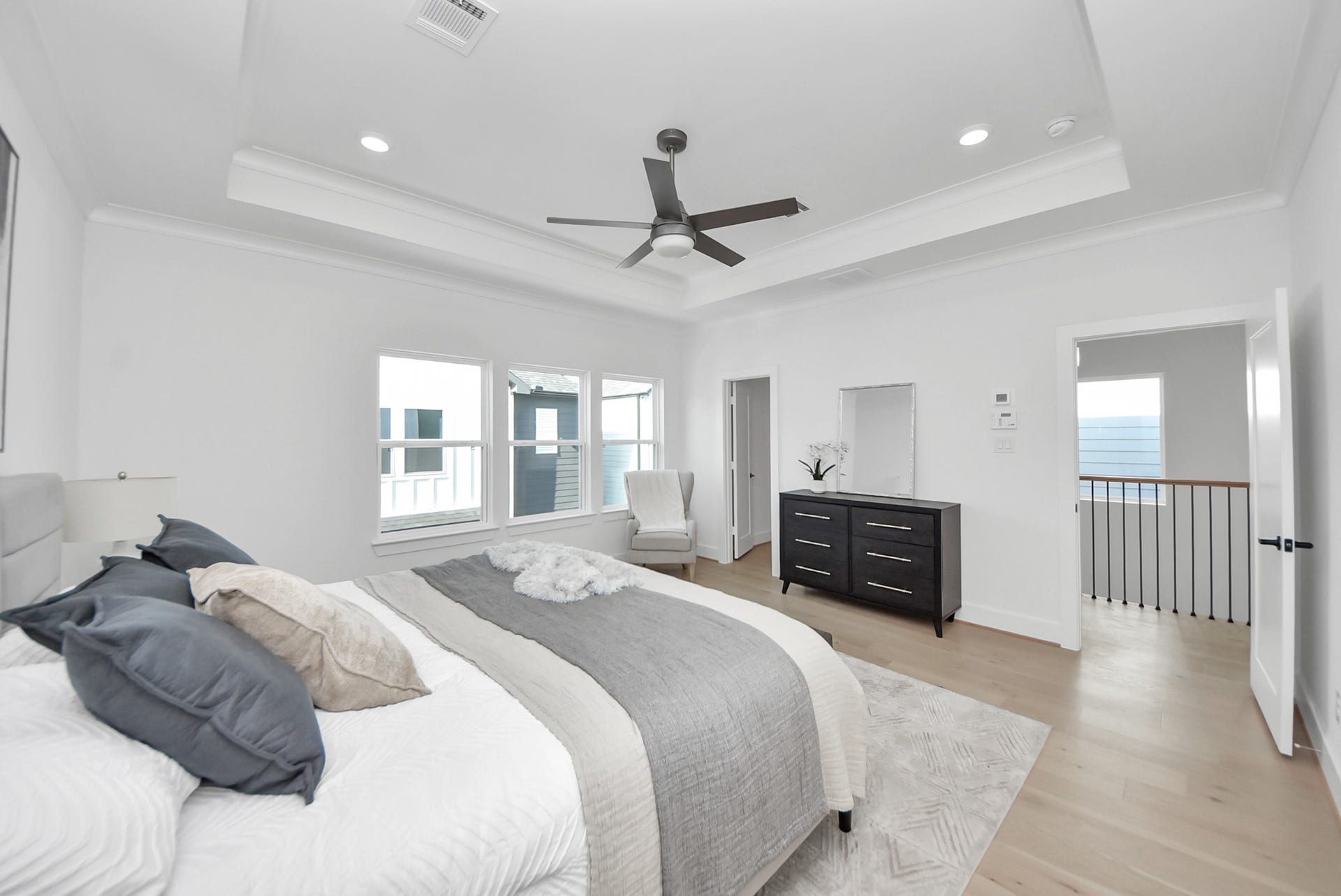 923 Fisher Street, #E, Houston, TX 77018 The primary bedroom impresses with a tray ceiling, recessed lighting, and a sleek ceiling fan. Large windows provide ample sunlight, and the comfortable bed with chic bedding, paired with a dresser and mirror, creates an open and inviting space that connects seamlessly to additional rooms.