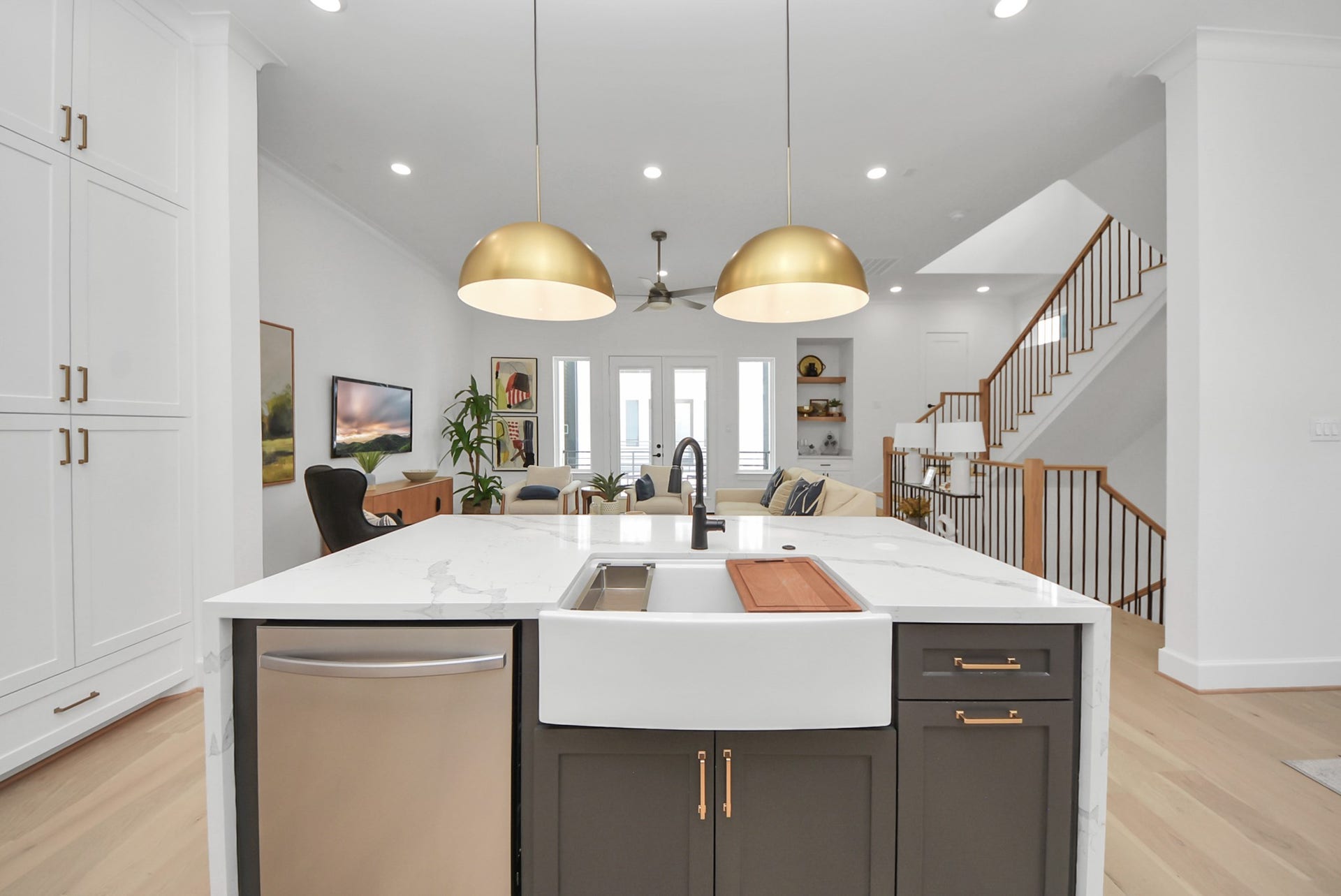 923 Fisher Street, #E, Houston, TX 77018 Enjoy this modern open-concept kitchen and living area, featuring a large island with a farmhouse sink, stainless steel appliances, and gold pendant lighting. The space opens to a cozy living room with natural light, a ceiling fan, and stylish dcor, while a wooden staircase adds warmth and architectural interest.