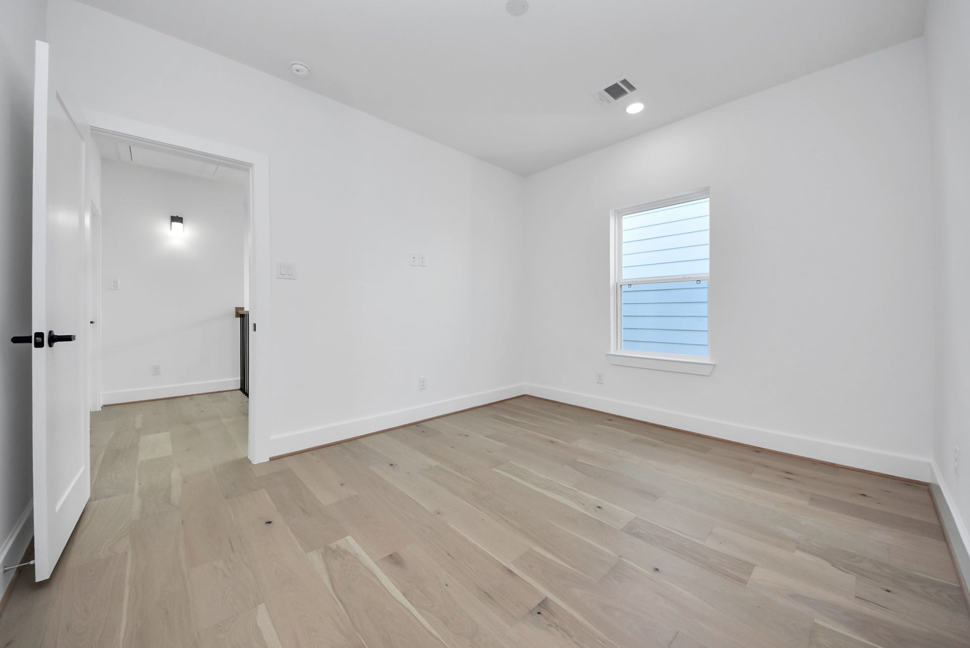 923 Fisher Street, #E, Houston, TX 77018 This room offers a minimalist design with light wood floors, white walls, and recessed lighting. A single window lets in natural light, while a white door connects to a well-lit hallway, offering versatility for a bedroom, office, or other use.