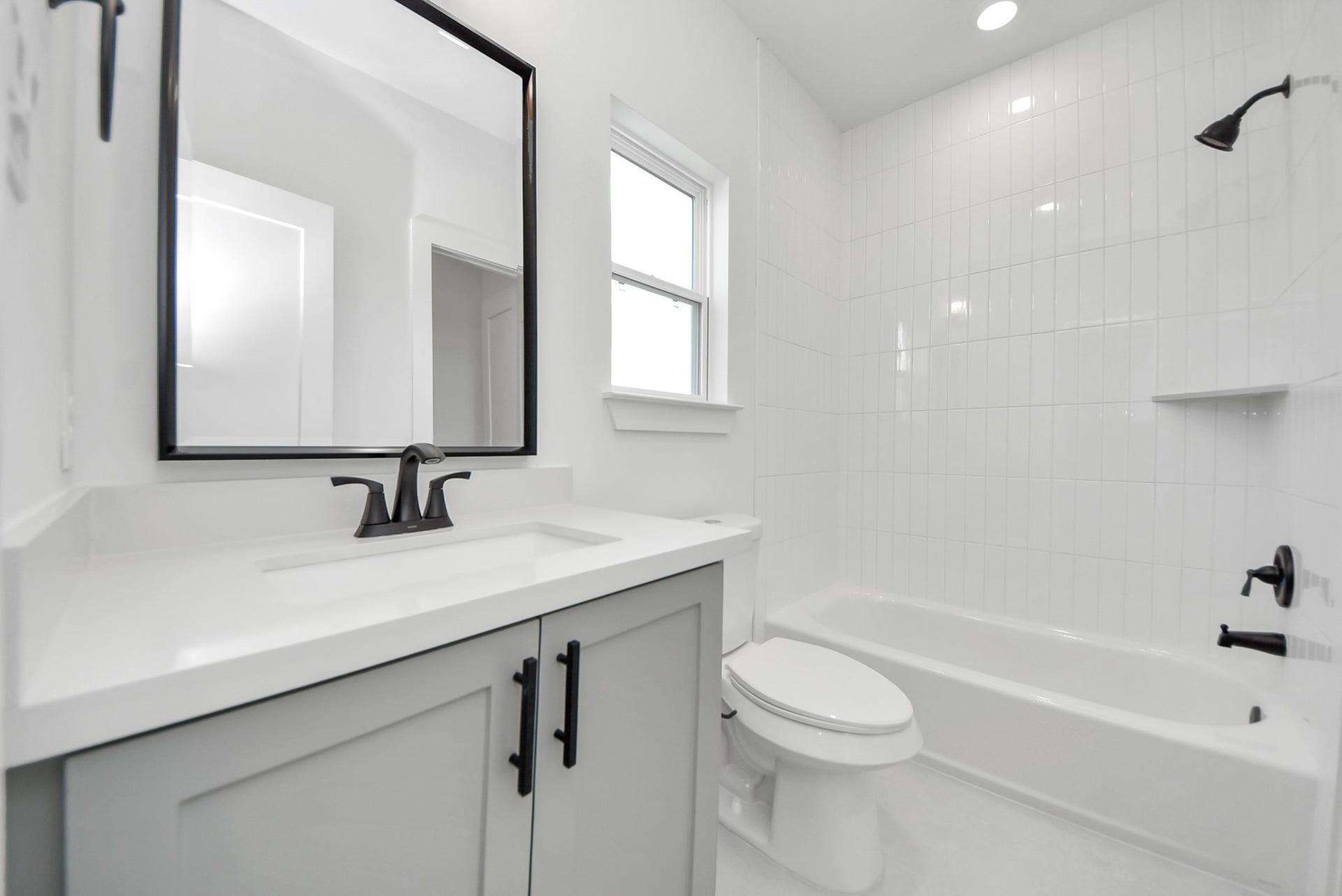 923 Fisher Street, #E, Houston, TX 77018 This modern bathroom features a sleek vanity with a large mirror, a bathtub surrounded by white tile, and a window that brings in natural light, creating a bright and welcoming atmosphere.