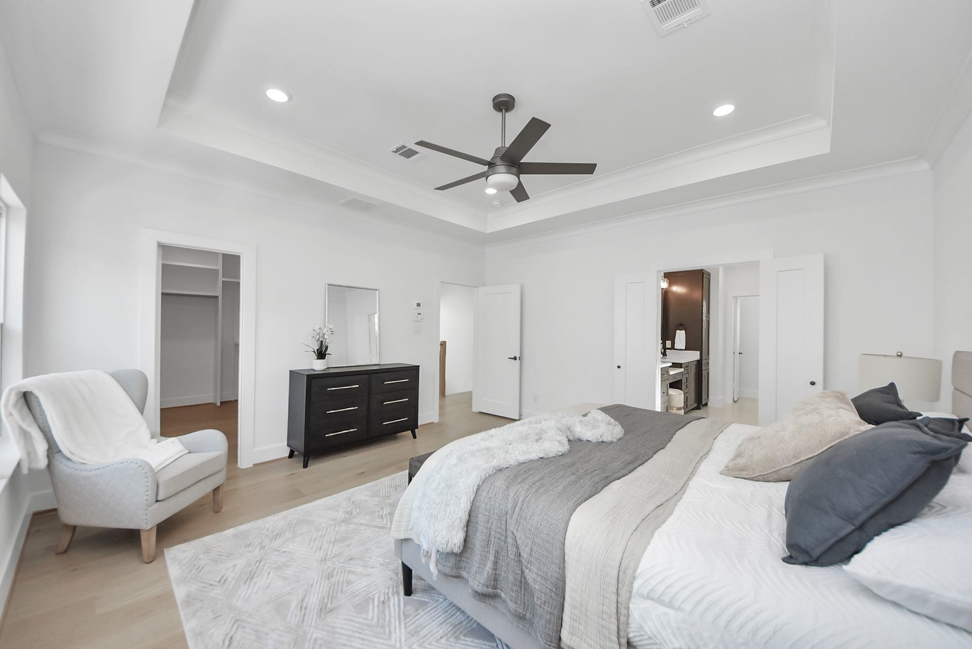 923 Fisher Street, #E, Houston, TX 77018 This modern bedroom features a neutral palette, recessed lighting, and a contemporary ceiling fan. A cozy sitting area, dresser, and access to a walk-in closet and en-suite bathroom make this room both functional and stylish, offering a perfect personal retreat.