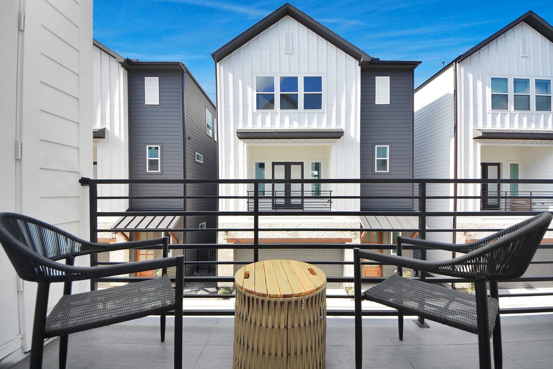 923 Fisher Street, #E, Houston, TX 77018 Step out onto this modern balcony with sleek black railings and outdoor seating, overlooking contemporary townhouses with contrasting exteriors. The space is perfect for relaxing and enjoying neighborhood views under a clear blue sky.