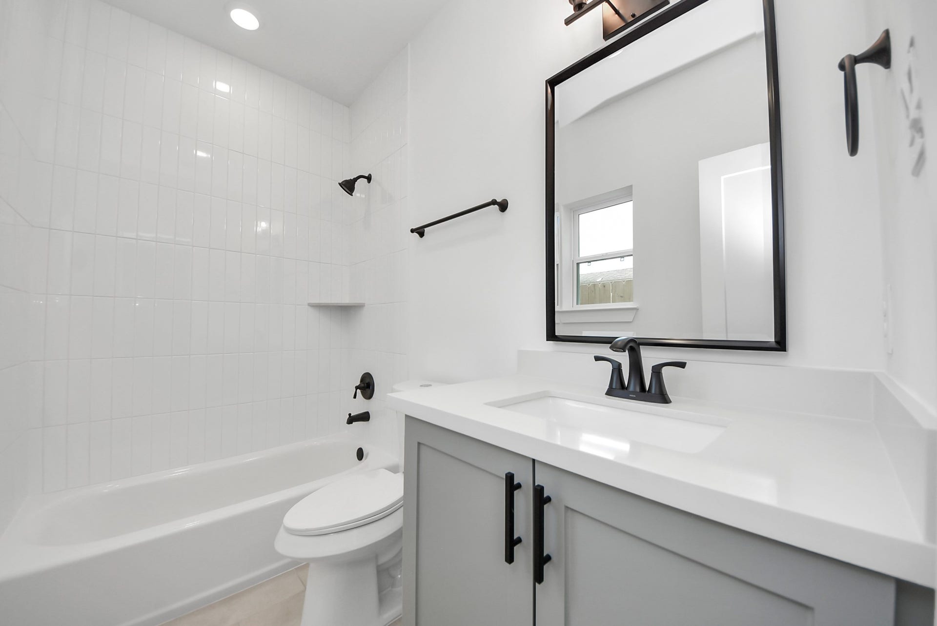 923 Fisher Street, #E, Houston, TX 77018 This bathroom features a clean, modern design with white tile, a bathtub-shower combination, and a sleek vanity with a large mirror. Black fixtures add a stylish contrast, creating a bright and contemporary space.