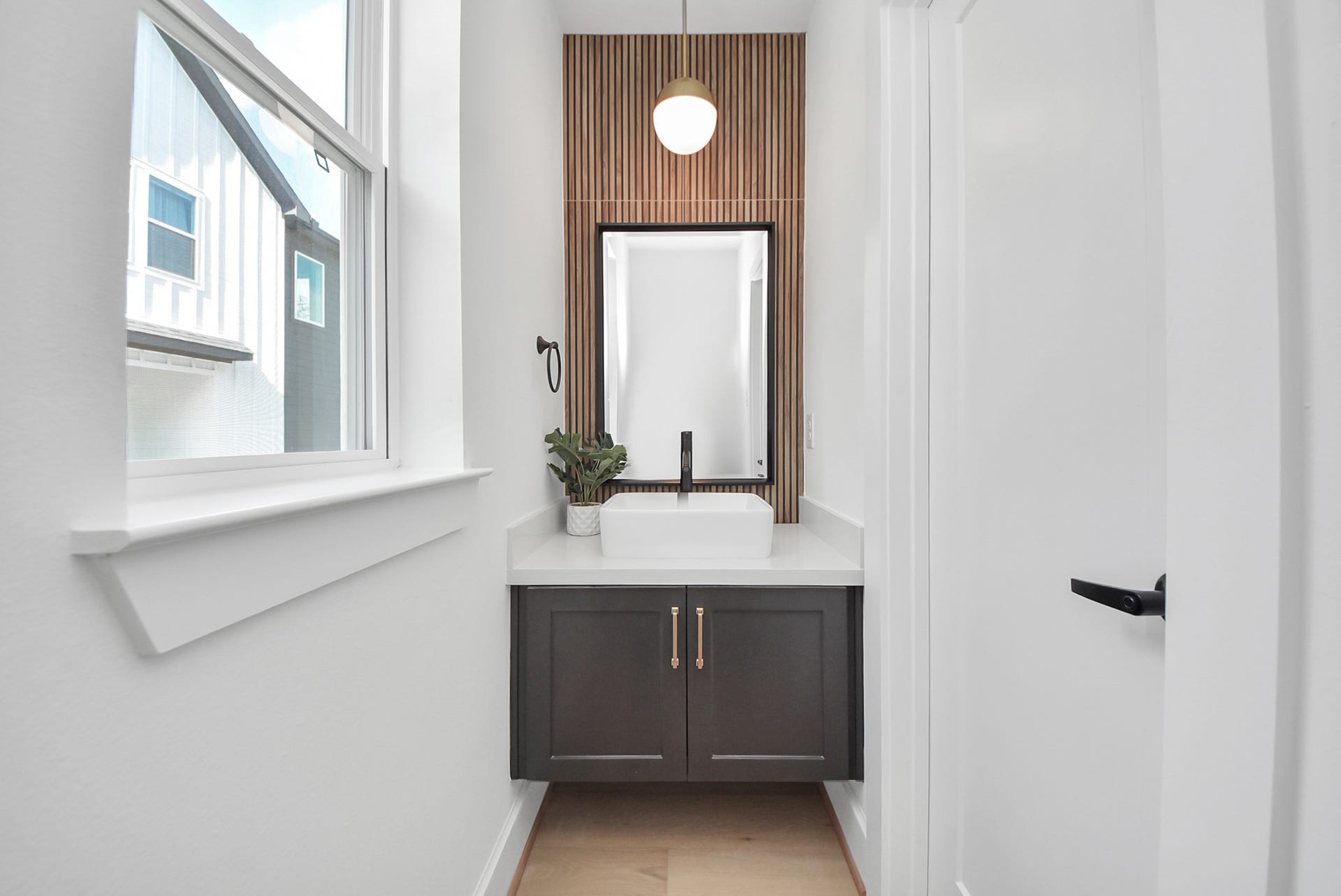 923 Fisher Street, #E, Houston, TX 77018 Step into this modern, compact bathroom featuring a sleek vanity with a white countertop and vessel sink. A stylish pendant light hangs above a wood-accent wall, while natural light from the window brightens the space, creating a clean and inviting design.