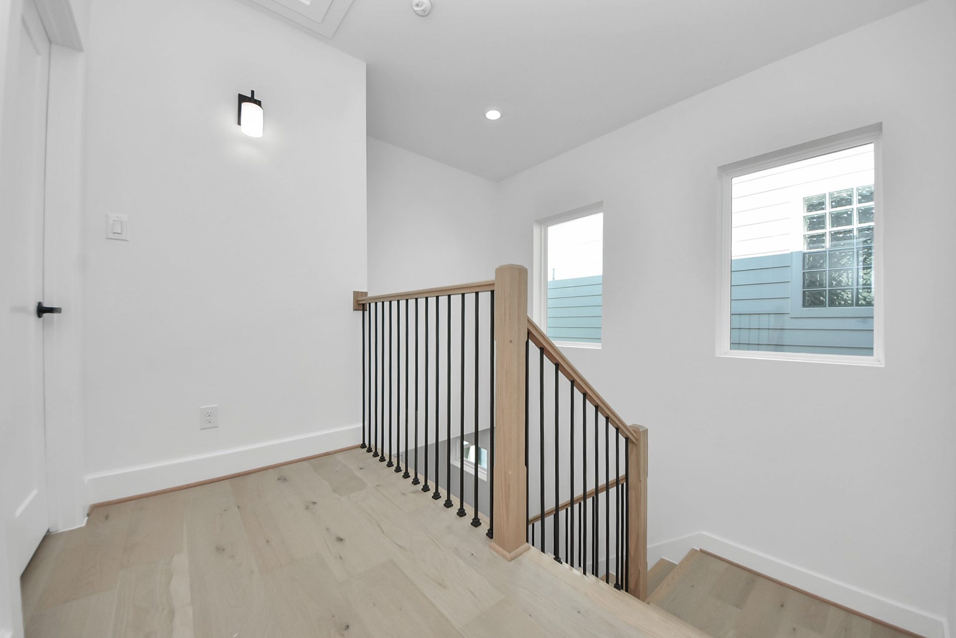 923 Fisher Street, #E, Houston, TX 77018 This bright, modern stairway landing features light wood flooring and a sleek black metal railing. Two large windows flood the space with natural light, creating an open and inviting atmosphere.
