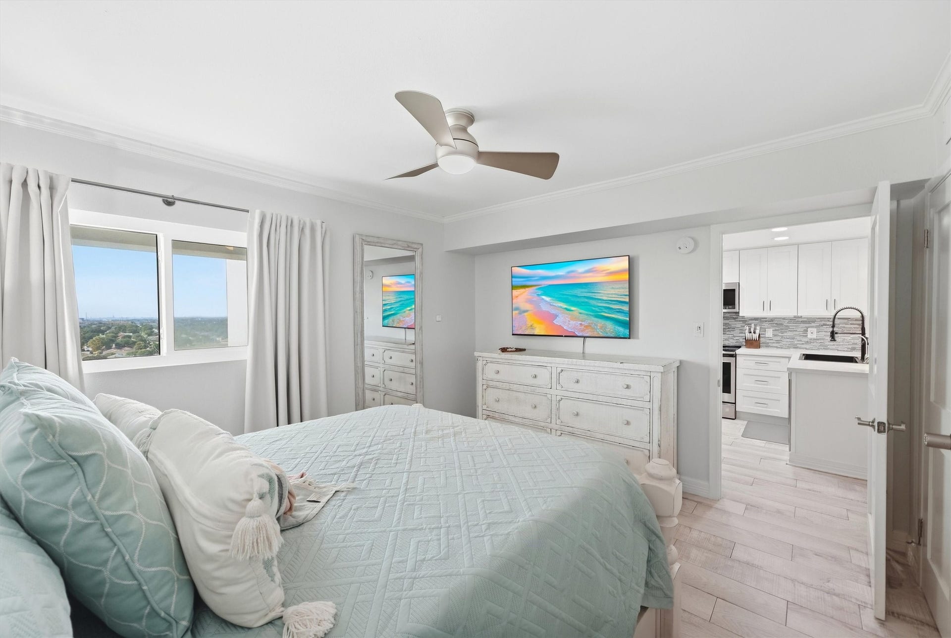 7310 Seawall Boulevard, #1105, Galveston, TX 77551 Bright and airy bedroom with large window views, a cozy bed with soft pastel linens, and modern ceiling fan. The room opens into a sleek, white kitchen, adding a sense of spaciousness and flow.
