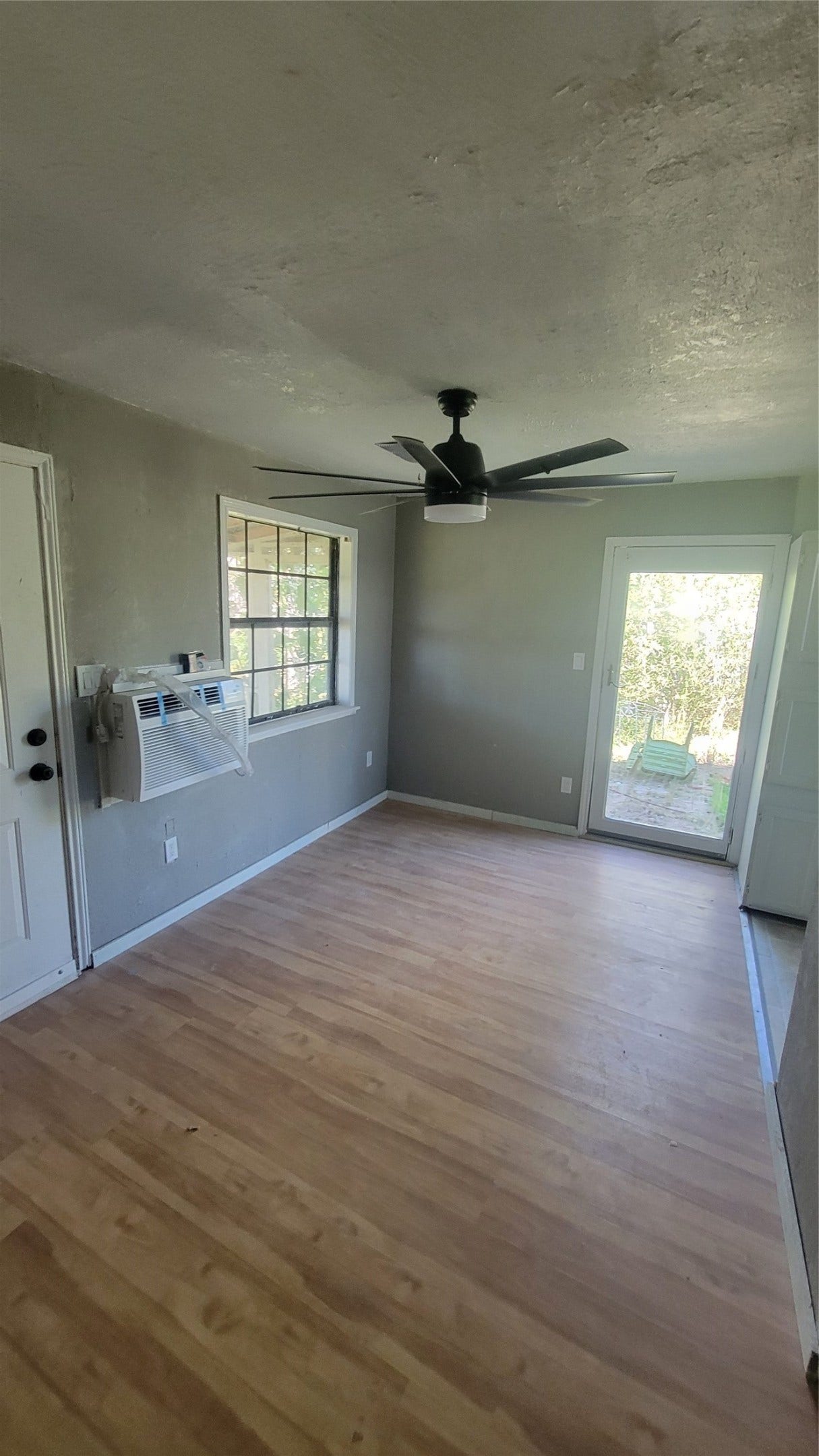 12018 Sandpiper Road, Angleton, TX 77515 This room features hardwood floors, a ceiling fan, and a wall-mounted air conditioning unit. It has a window for natural light and a door leading to an outdoor area. Ideal for a cozy living space or office.
