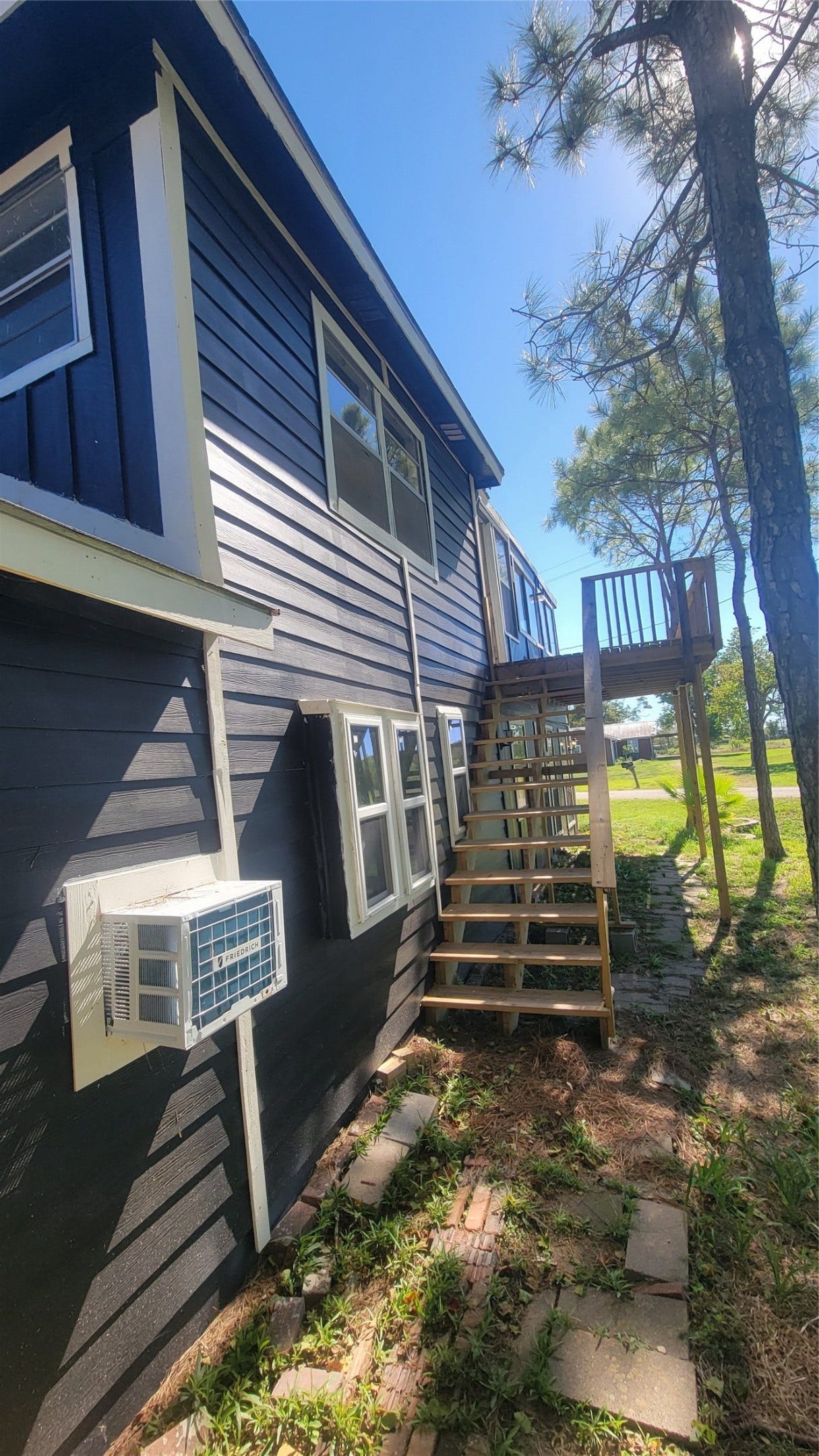 12018 Sandpiper Road, Angleton, TX 77515 This photo showcases the side of a two-story home with dark siding, featuring a staircase leading to the upper level. There's an air conditioning unit on the lower wall, surrounded by a path of stepping stones and some greenery. The setting is bright and sunny, with a few trees nearby.
