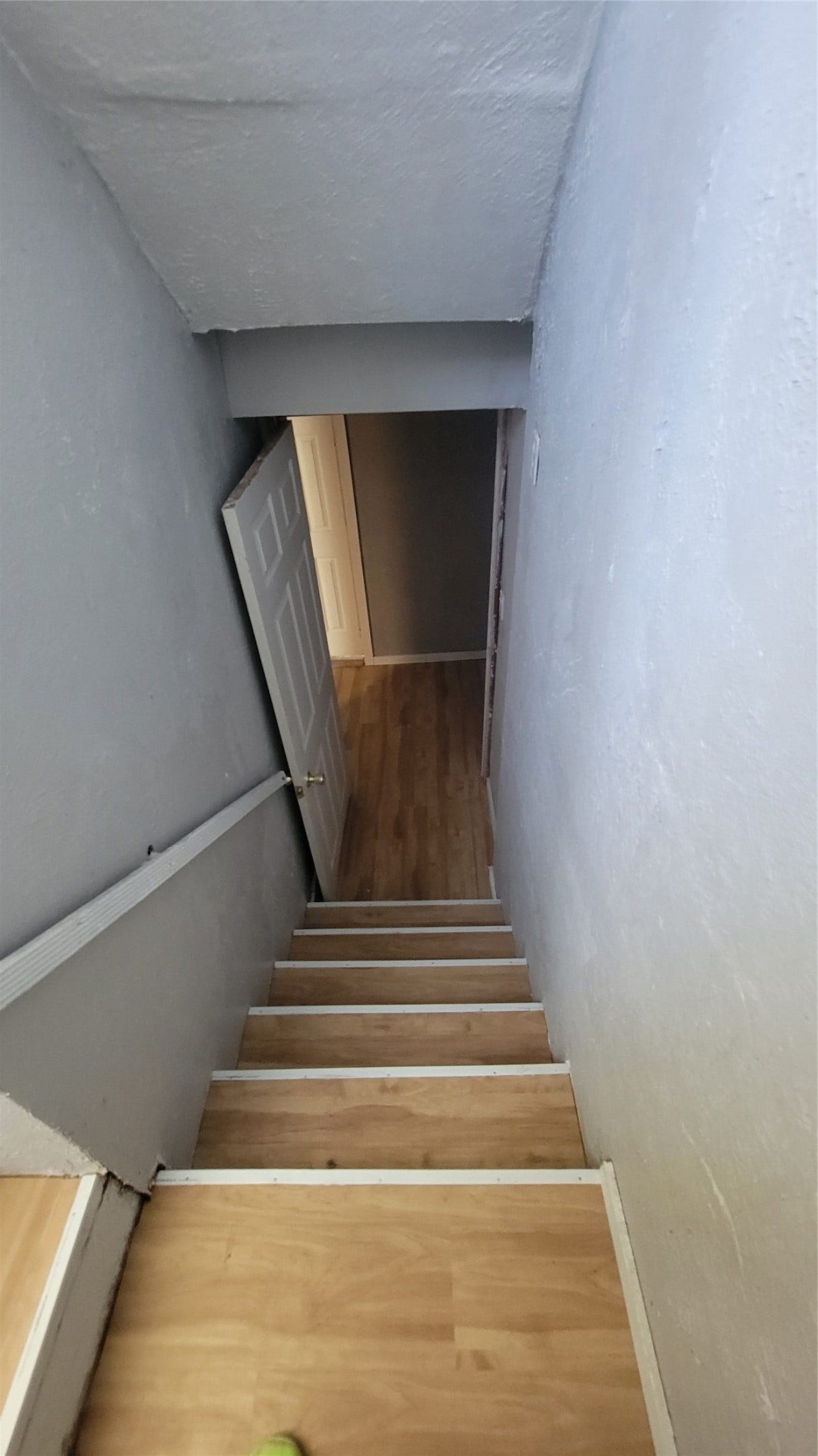 12018 Sandpiper Road, Angleton, TX 77515 This photo shows a narrow staircase with wooden steps leading down to a lower level. The walls are painted light gray, and there's a white door at the bottom opening into a room with similar flooring.