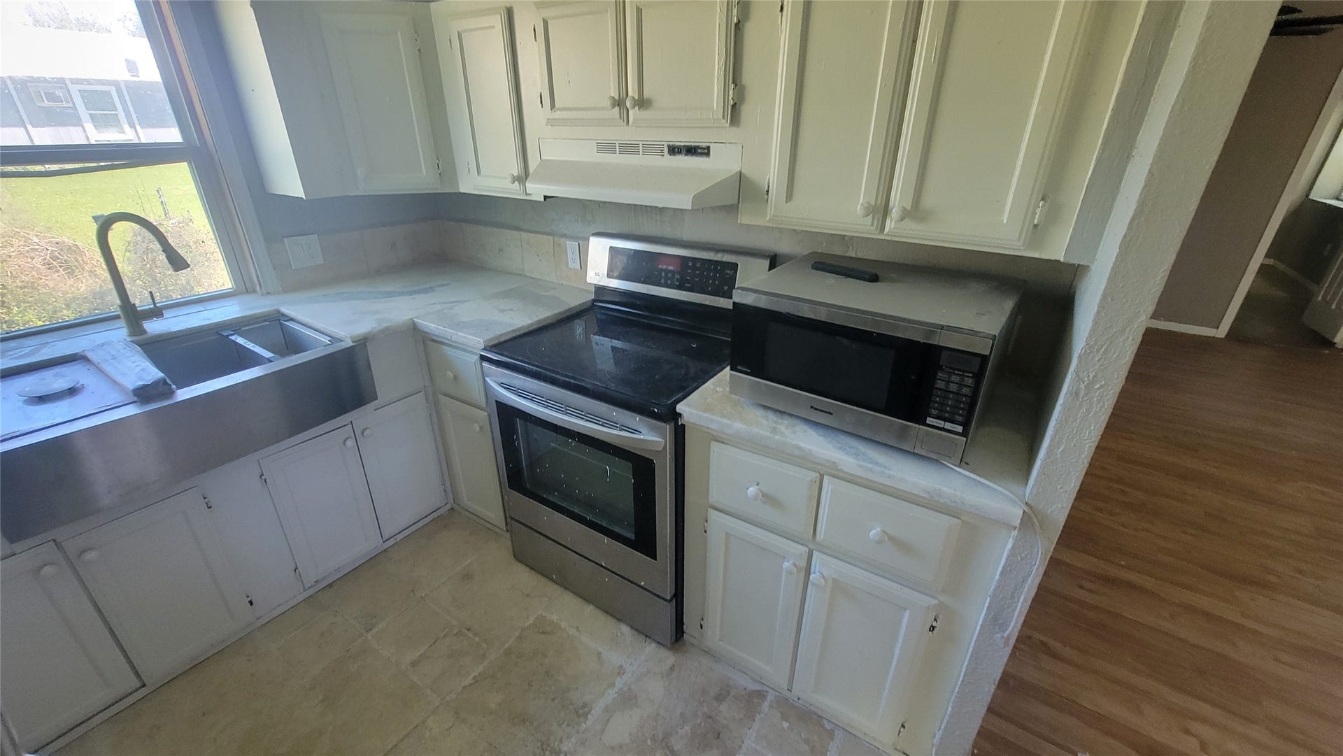 12018 Sandpiper Road, Angleton, TX 77515 This kitchen features white cabinetry, a stainless steel farmhouse sink, and a modern electric stove with a microwave. The space is brightened by a large window and has a tiled floor with adjacent wood flooring.