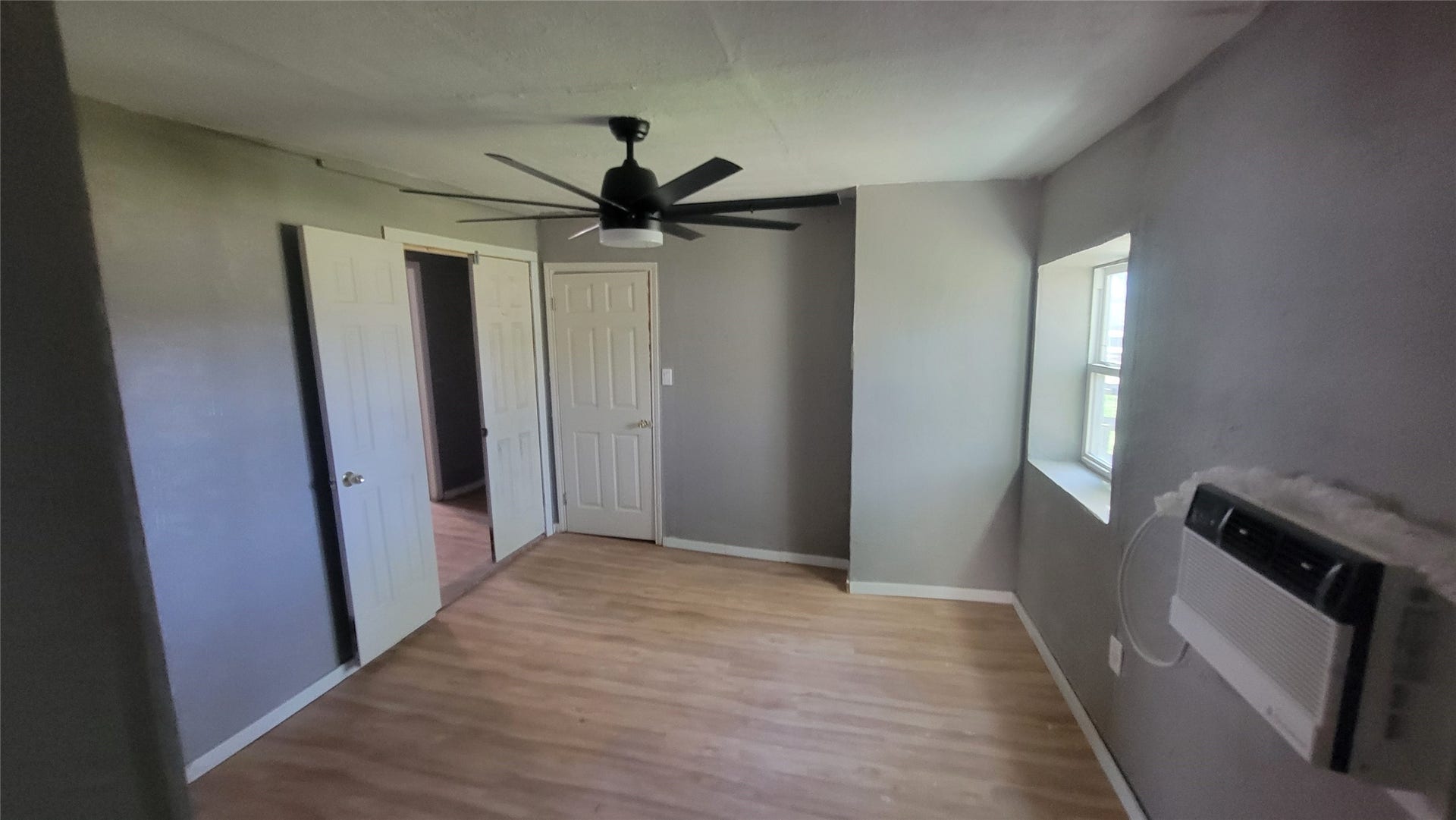 12018 Sandpiper Road, Angleton, TX 77515 This room features light wood flooring, neutral gray walls, a window with ample natural light, a ceiling fan, and wall-mounted air conditioning. It's a cozy space suitable for a variety of uses.