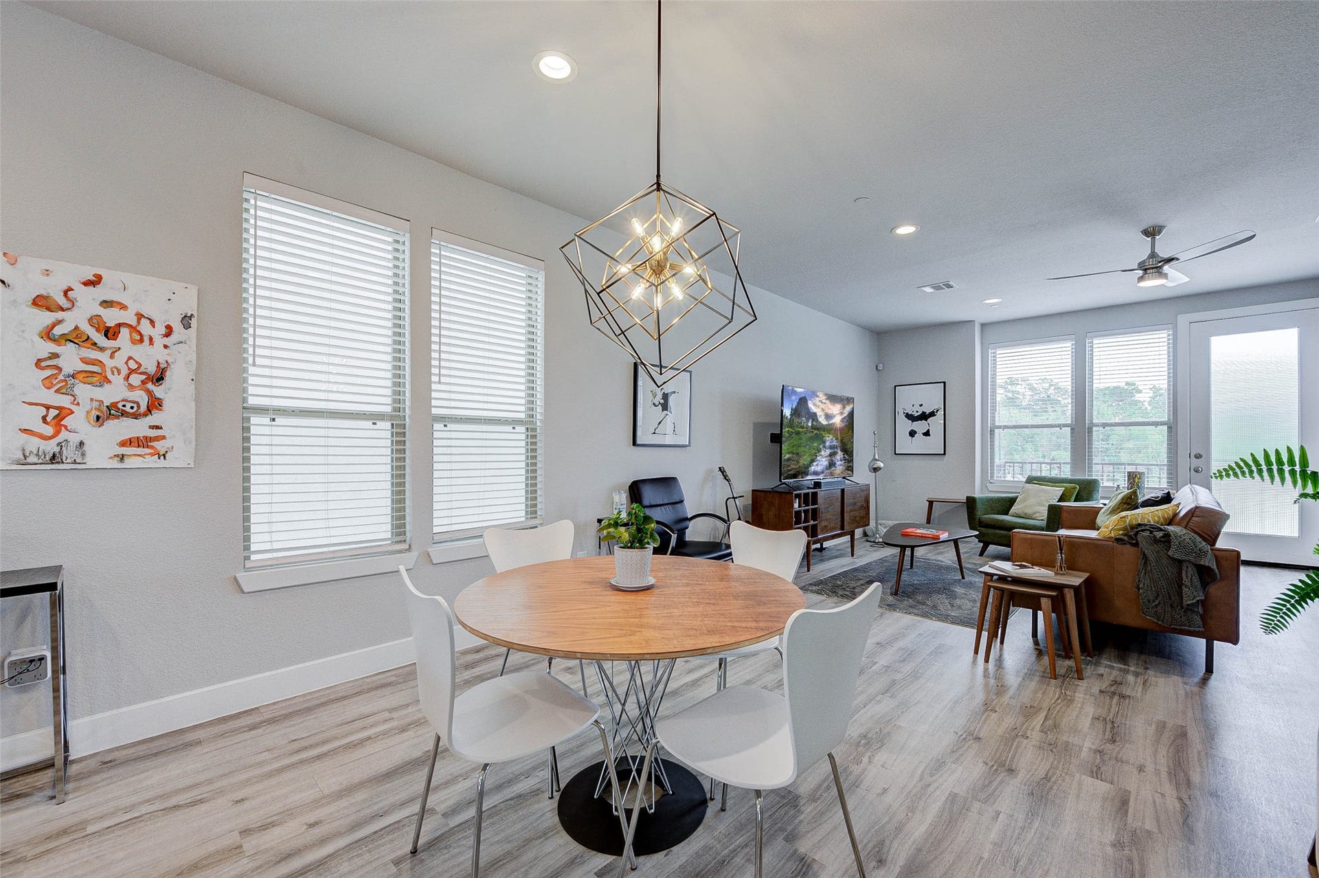 18732 Centro Main Street, Shenandoah, TX 77385 This photo showcases a modern, open-concept living and dining area with light wood flooring and ample natural light from large windows. The space features contemporary furniture, including a round dining table, stylish chairs, and a cozy seating area with a sofa and TV. A unique geometric light fixture adds a touch of elegance.