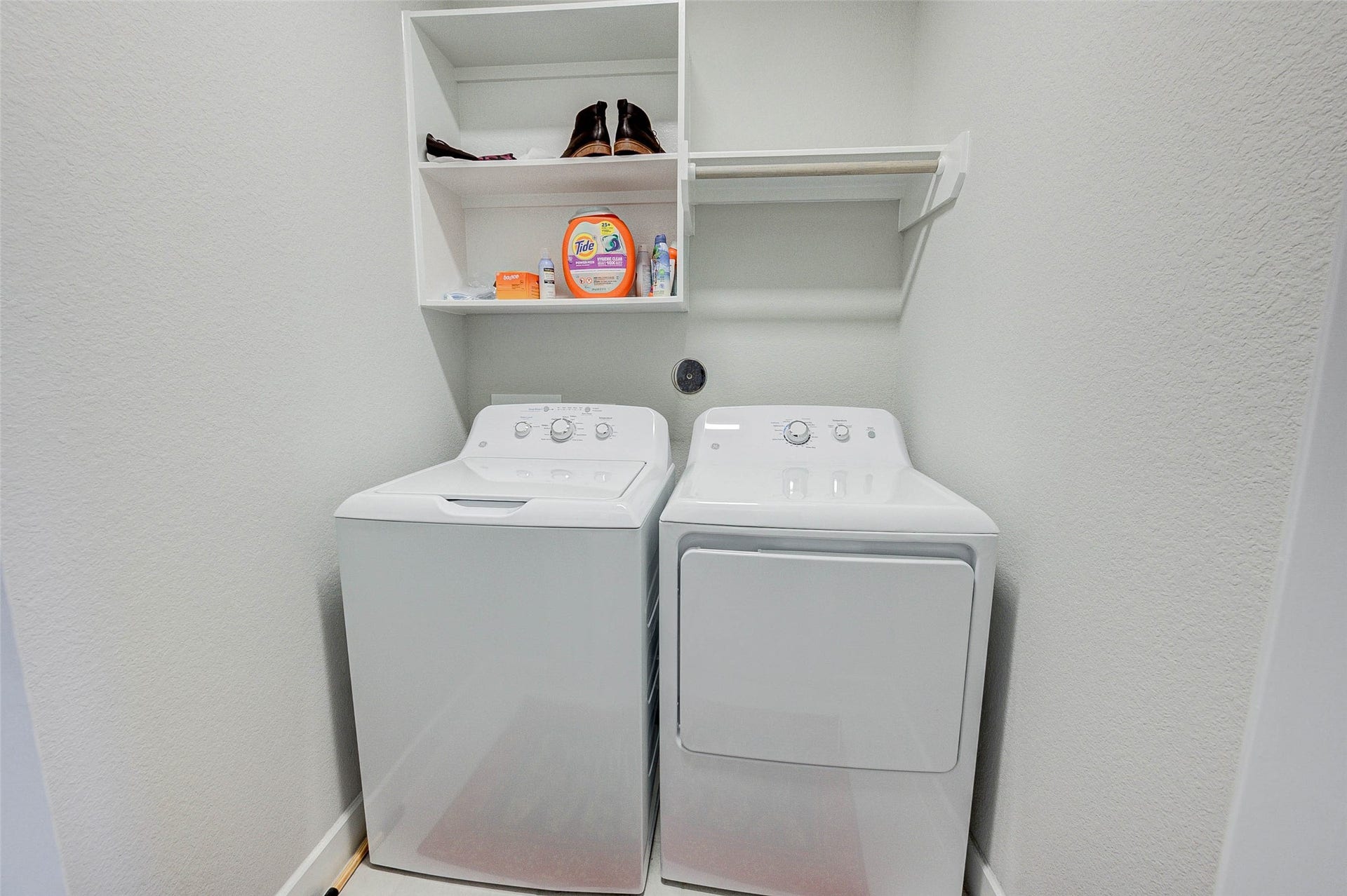 18732 Centro Main Street, Shenandoah, TX 77385 This photo shows a compact laundry area with a washer and dryer side by side. Above the appliances, there's shelving for storage, ideal for keeping detergents and laundry essentials organized. The space is clean and efficiently designed.