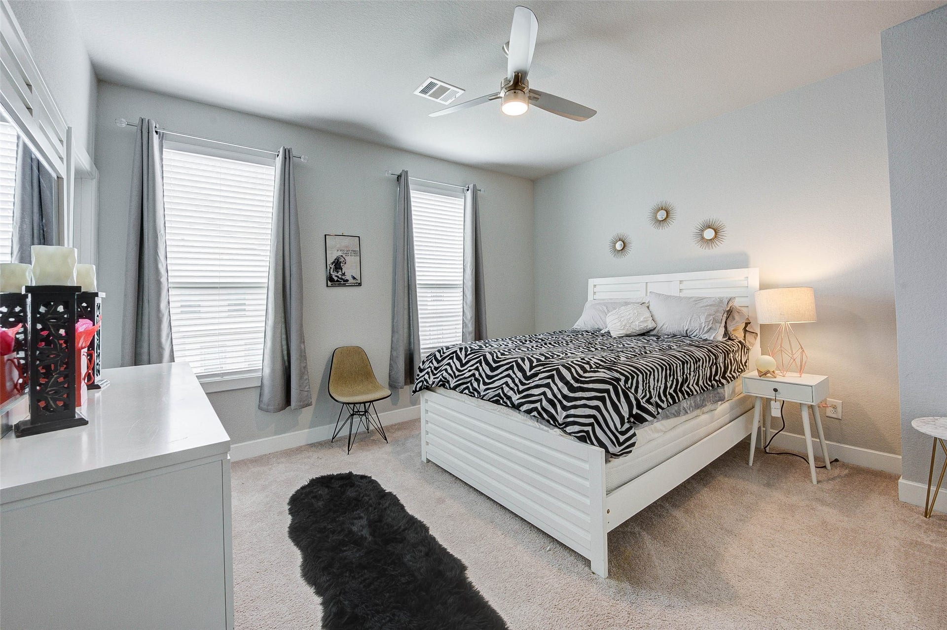 18732 Centro Main Street, Shenandoah, TX 77385 This bedroom features a cozy, modern design with soft carpet flooring and neutral walls. It includes a stylish bed with a zebra-patterned duvet, a ceiling fan, and ample natural light from two large windows with gray curtains. A chic dresser and nightstand, complemented by decorative wall accents, complete the room.