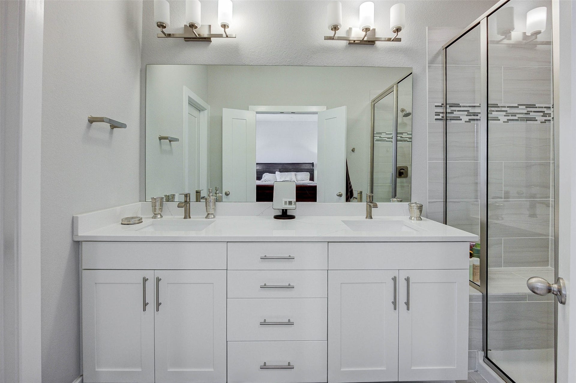 18732 Centro Main Street, Shenandoah, TX 77385 This modern bathroom features a spacious double vanity with sleek white cabinetry and dual sinks. It includes a large mirror and elegant lighting fixtures, with a glass-enclosed shower showcasing stylish tile work.