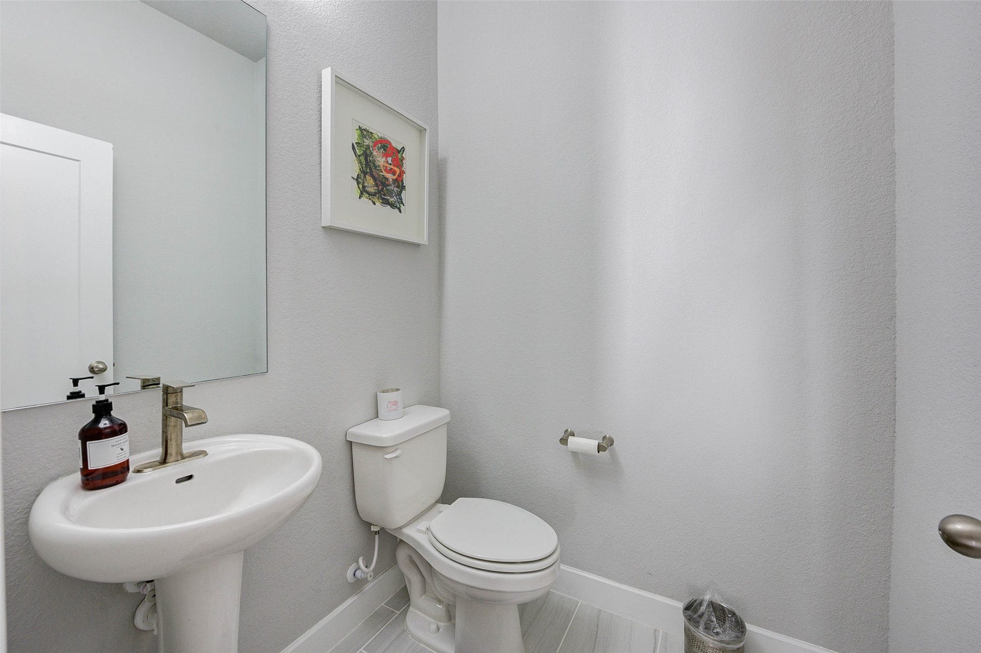 18732 Centro Main Street, Shenandoah, TX 77385 This is a modern, compact half-bathroom featuring a pedestal sink, a toilet, and neutral-toned walls. It includes a large mirror, a small wall art piece, and has a clean, minimalistic design.