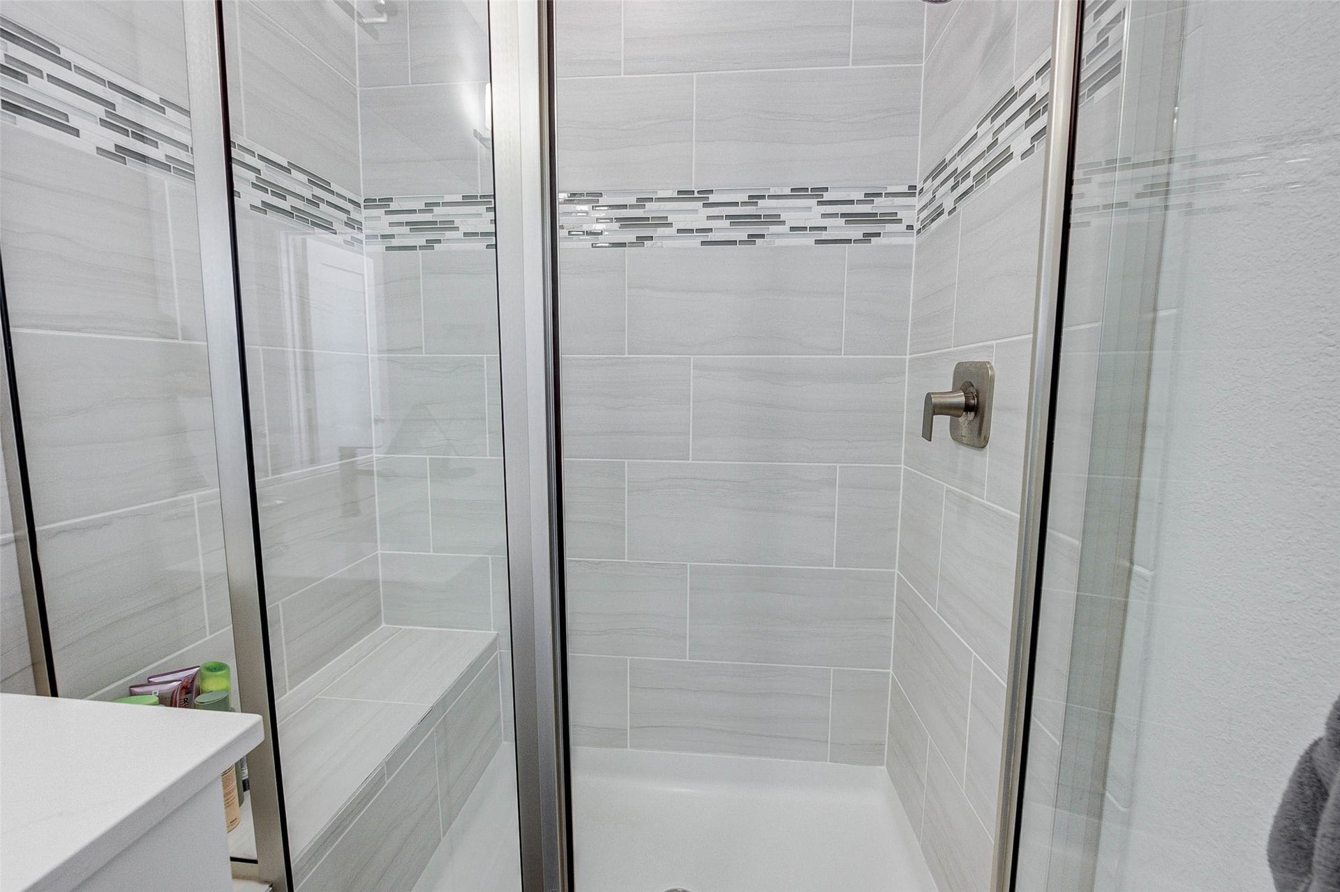 18732 Centro Main Street, Shenandoah, TX 77385 This photo shows a modern shower with sleek glass doors and a built-in bench. The walls are lined with light gray tiles accented by a decorative band of smaller, darker tiles, creating a stylish and contemporary look.