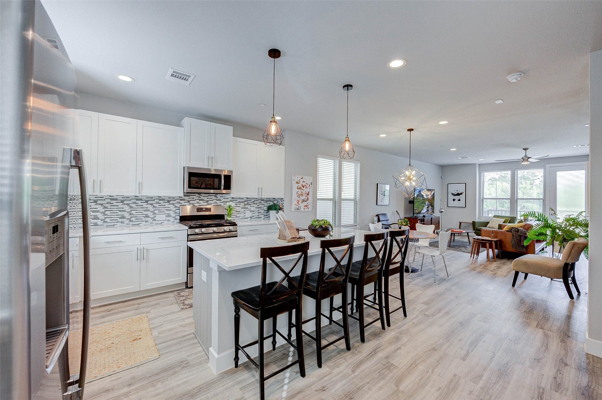18732 Centro Main Street, Shenandoah, TX 77385 Modern open-concept kitchen and living area with sleek white cabinetry, stainless steel appliances, and stylish pendant lighting. The space features a functional island with seating, light wood flooring, and ample natural light from large windows, creating a welcoming atmosphere.