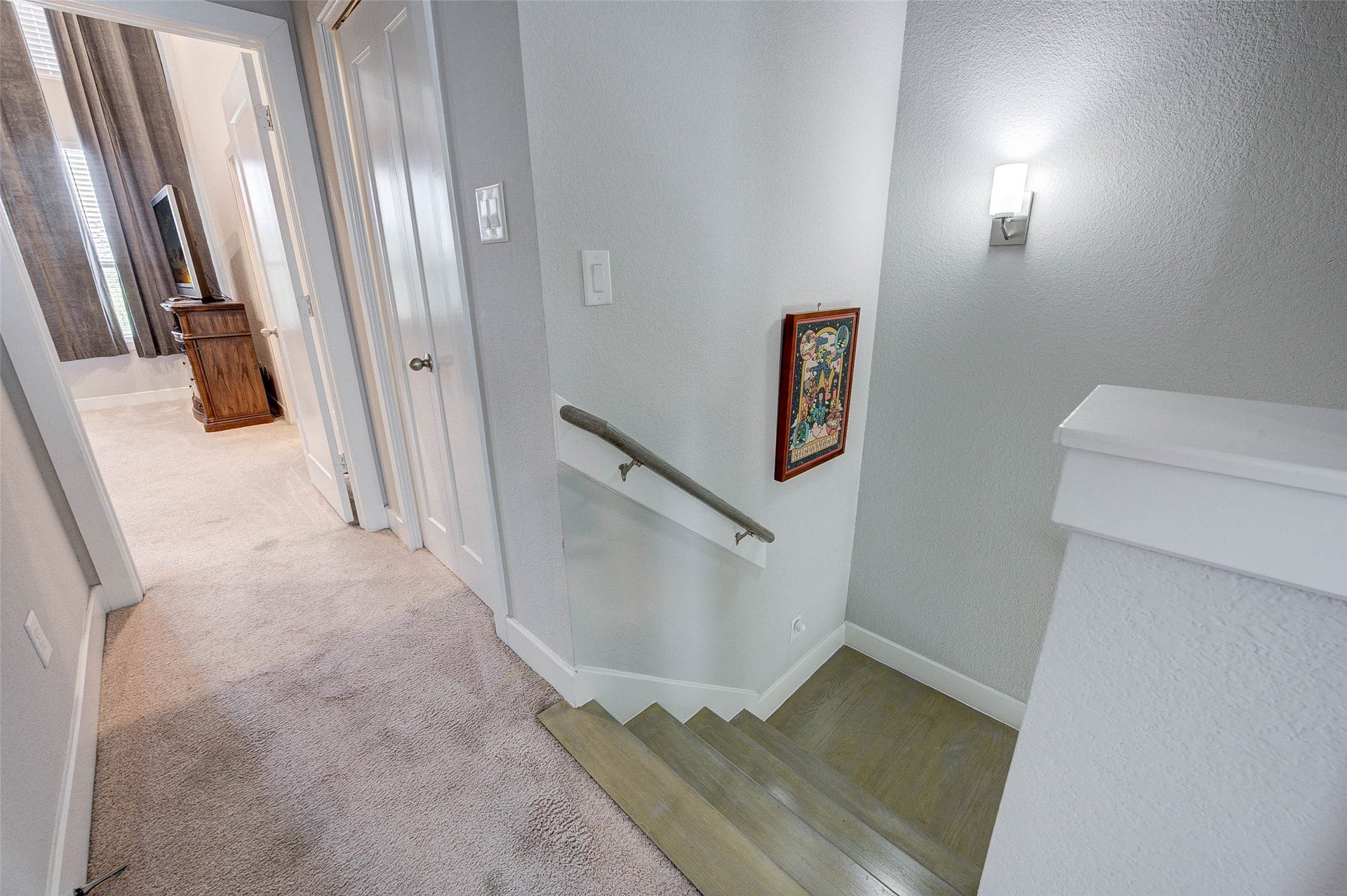 18732 Centro Main Street, Shenandoah, TX 77385 This photo shows a bright, carpeted hallway leading to a bedroom with large windows and a TV. It features a well-lit stairway with a wooden handrail and a decorative wall piece.